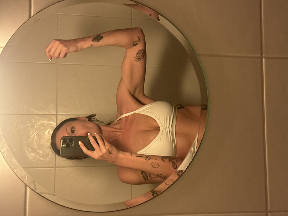 A woman stands sideways in front of a round mirror, holding a smartphone to take a selfie. Her body is angled slightly towards the viewer, showcasing her arm flexed at the bicep. She wears a light-colored top and displays multiple tattoos on her arms and torso. The background features tiled walls. The lighting appears muted, possibly from a bathroom or similar setting.