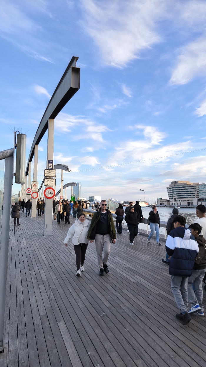 The image captures a vibrant scene on a wide wooden pier or boardwalk under a bright blue sky with scattered, wispy white clouds. Numerous people are walking along the pier, suggesting a public recreational area or a busy thoroughfare.
In the immediate foreground, a couple is walking towards the viewer, holding hands. The woman on the left is wearing a white puffy jacket, dark pants, and white sneakers. The man beside her is wearing a dark (possibly olive-green) jacket, light grey sweatpants, dark shoes, and sunglasses. To their right and slightly behind them, a younger individual in a blue and white puffer jacket walks alongside another person in a brown jacket. The wooden planks of the pier are laid lengthwise, exhibiting a weathered grey-brown tone.
Along the left side of the pier, a series of modern architectural elements consisting of grey metal beams and vertical supports create an overhead structure that extends into the distance. Attached to these supports are several signs. Prominently visible are two circular red signs with white backgrounds and black symbols: one depicts a crossed-out bicycle, and the other a crossed-out moped or scooter, indicating that these modes of transport are prohibited. Above these, a rectangular sign contains smaller text, likely detailing further regulations in a foreign language (possibly Spanish or Catalan). Anti-bird spikes are visible on the upper edges of some of the metal beams.
In the mid-ground and background, more people are seen walking in various directions, some individually and others in groups. To the right of the pier, a calm body of water (likely a harbor or bay) stretches towards a distant cityscape. A distinctive modern building with a curved, multi-story facade and numerous windows stands out on the far bank. Several boats or ships are partially visible in the water. Two seagulls are captured in flight: one in the mid-ground over the water, and another higher in the sky.
The weather appears clear and pleasant, with ample natural light suggesting it is daytime, possibly late afternoon given the quality of the light. Subtle, linear cloud patterns, possibly contrails, are faintly visible high in the sky. The overall atmosphere is lively and inviting, characteristic of a popular urban waterfront.