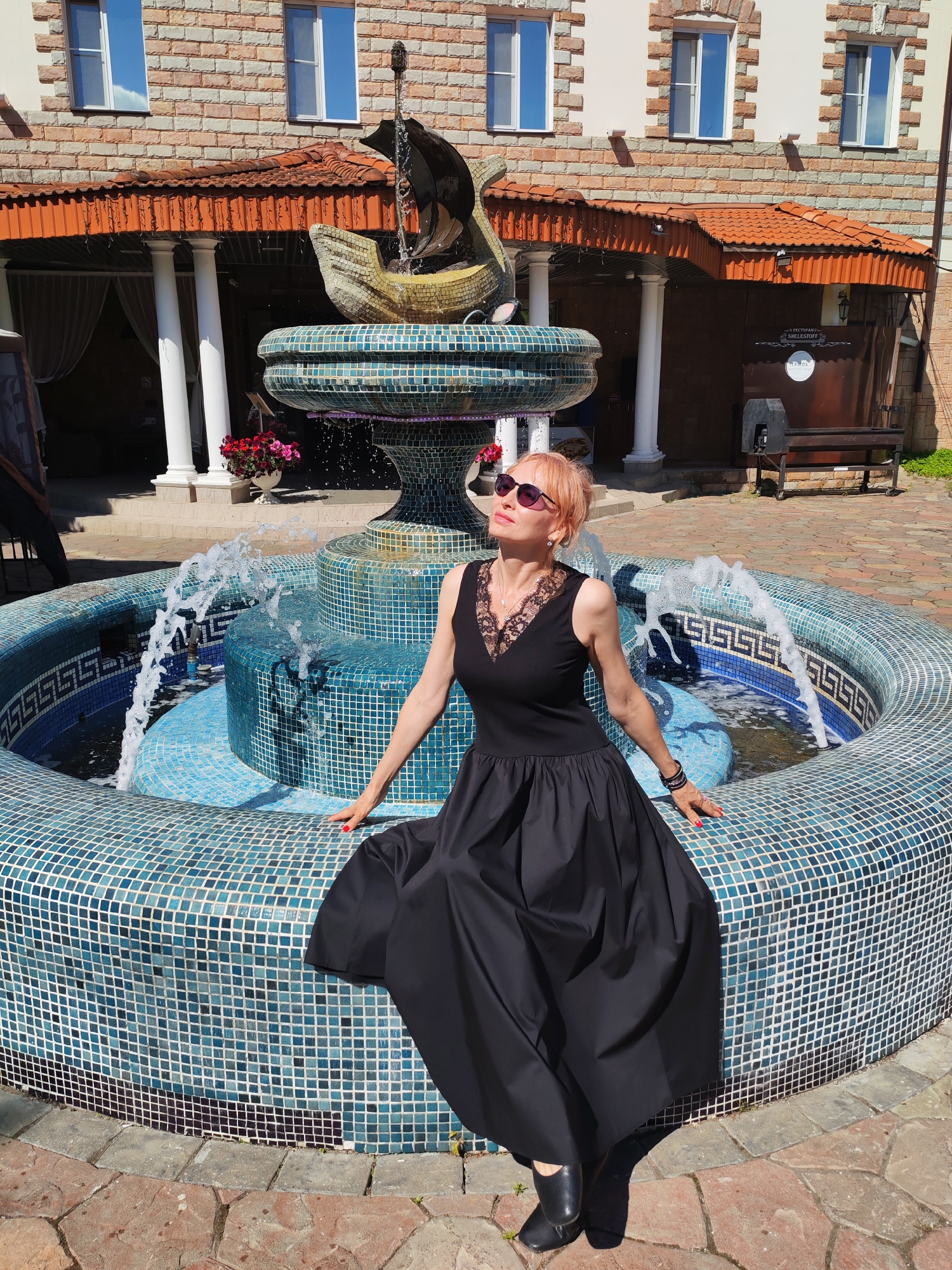 A woman with short, pink hair and sunglasses poses by a tiled fountain in an outdoor courtyard. She is wearing a black dress with a lace V-neck and black shoes. The fountain has a mosaic of blue and white tiles and features a statue of a ship at its center. In the background, a brick building with arched doorways and tiled roofs is visible. The courtyard is paved with stone. The scene is located at the restaurant "Shelestoff" in Kostroma, Russia.