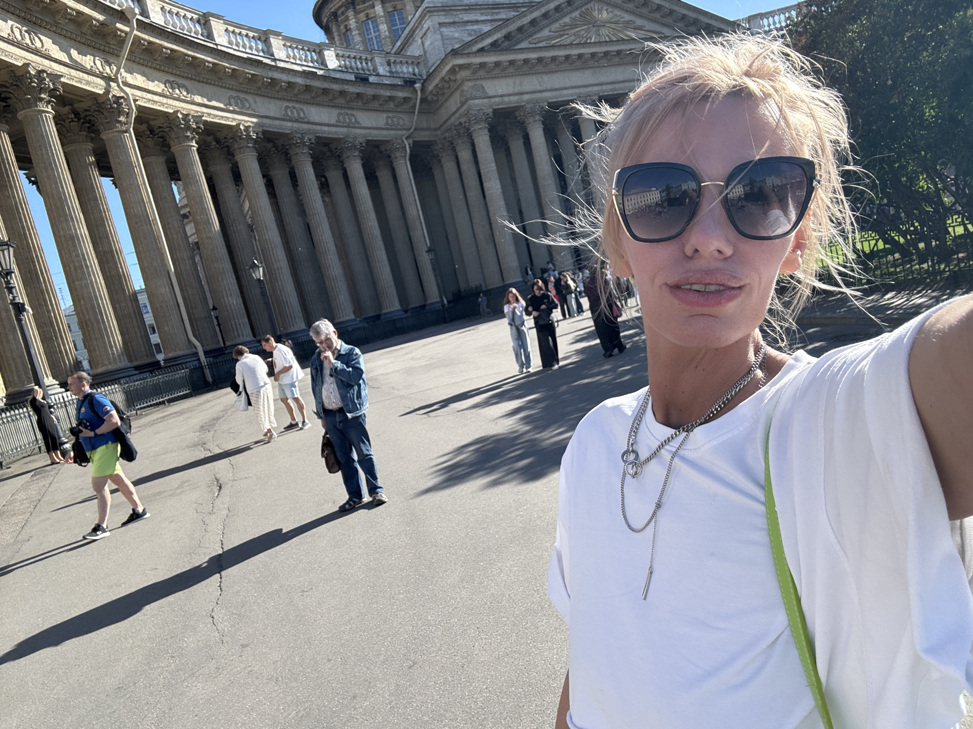 This media file captures a selfie taken by a woman with blonde hair and sunglasses, standing in front of a grand, colonnaded building. The building appears to be a historical landmark with a large dome and ornate architectural details. The woman is wearing a white t-shirt and a silver necklace. Her expression is neutral.
Several other people are visible in the background, walking on a paved area. Some are dressed casually, while others appear to be tourists. The sun is shining brightly, casting long shadows, suggesting it is daytime. The overall scene is one of an outdoor public space, possibly a plaza or courtyard, in front of an important architectural structure. The presence of a camera in the selfie and the people strolling in the background suggest a leisurely or tourist activity. There is no visible text in the image. The location context provided identifies the setting as Saint Petersburg, Russia.