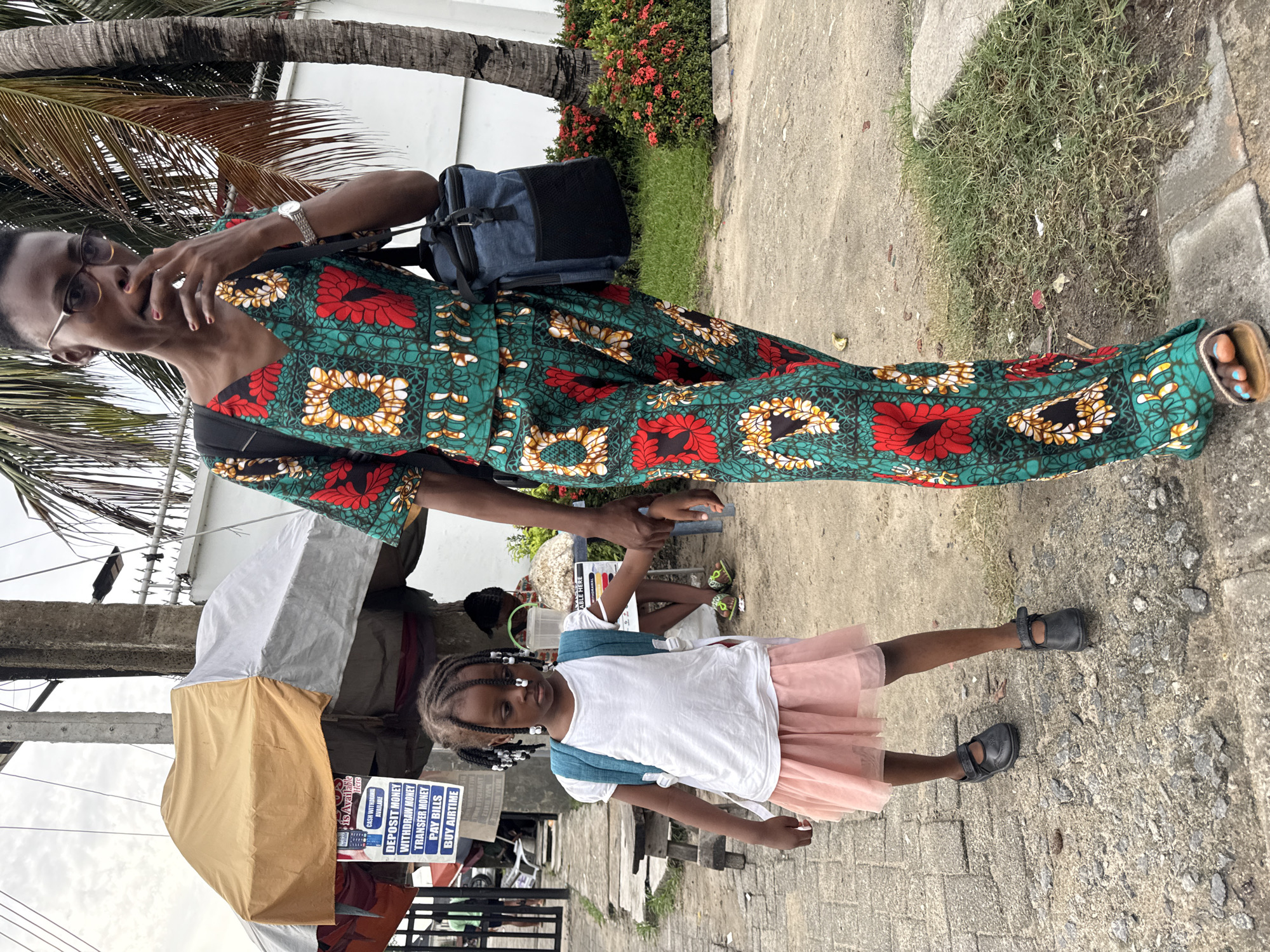 A full-length shot shows a woman and a young girl standing outdoors in what appears to be the city of Ado, Nigeria. The woman is wearing a colorful, long-sleeved jumpsuit with a green, red, and yellow floral pattern, glasses, and a wristwatch. She carries a blue bag and a black backpack, and holds the girl's hand. The girl wears a white t-shirt, a pink tutu, a blue backpack, and black shoes. They stand on a paved surface near a roadside business offering money transfer services, indicated by a sign. A partially obscured canopy covers the business. A palm tree and some greenery can be seen in the background.