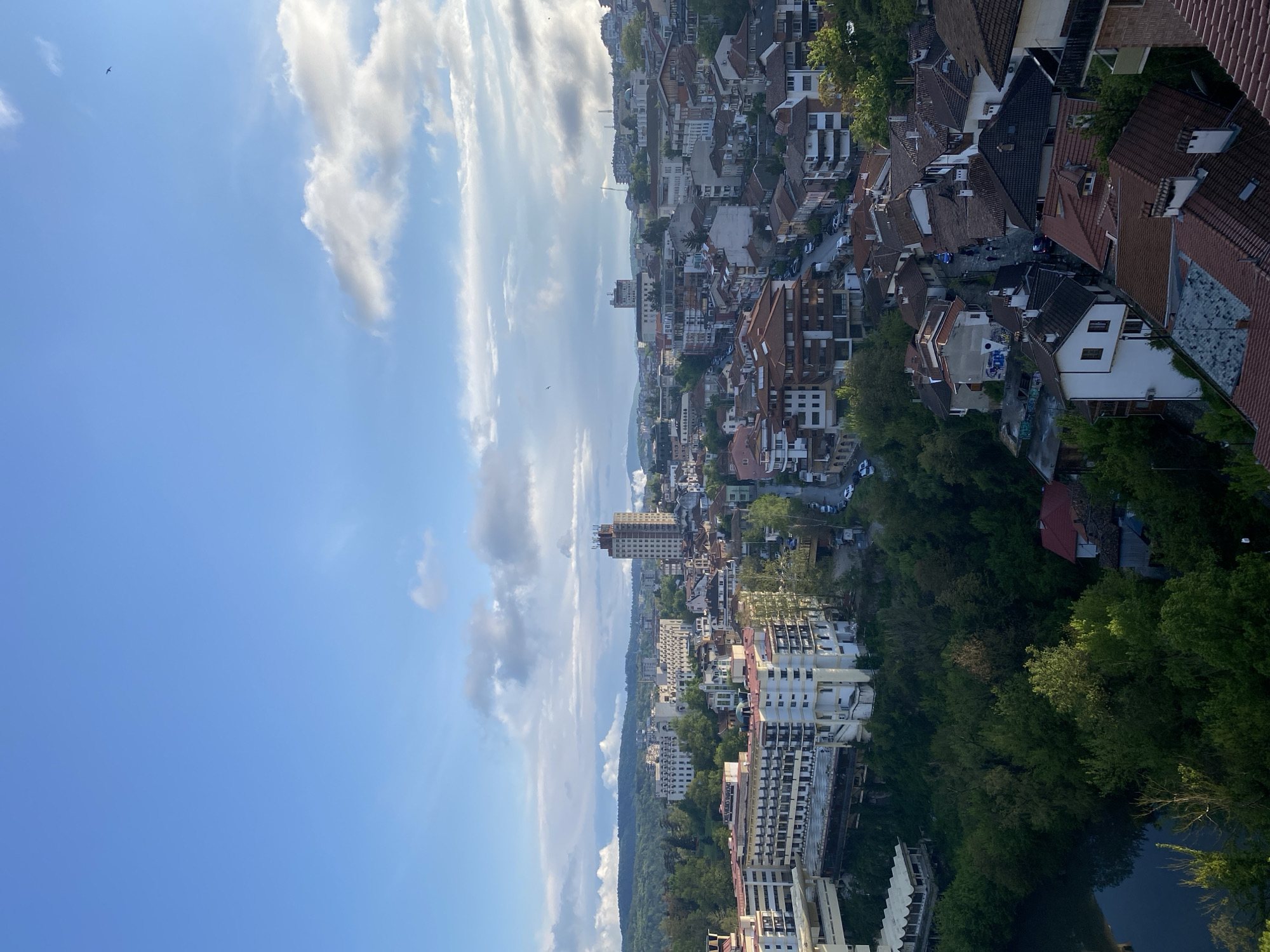 The image presents an aerial view of a city, possibly located in a hilly region. The sky is a mix of blue and white clouds, dominating the upper portion of the image. Below, a dense urban area unfolds with buildings of varying heights and architectural styles. A mix of residential houses with red-tiled roofs and taller, modern apartment buildings are visible. Patches of green foliage intersperse with the buildings, suggesting parks or tree-lined streets. In the distance, a mountain range is visible on the horizon. The overall impression is that of a densely populated urban environment nestled within a natural landscape.