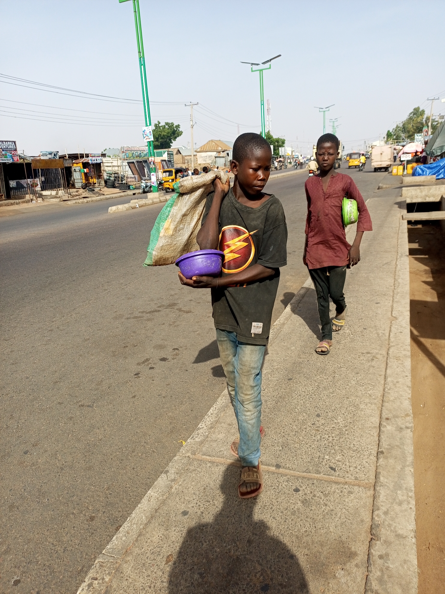 The image depicts two young boys walking along a street in Gombe, Nigeria. The boy in the foreground carries a sack over his shoulder and a purple bowl in his hand. He wears a t-shirt with a Flash logo and faded jeans. The other boy walks slightly behind him, carrying a green bowl. He is dressed in a maroon shirt and dark trousers. They are walking on a concrete sidewalk beside a busy road with shops and vehicles in the background. There are green street lights along the road.