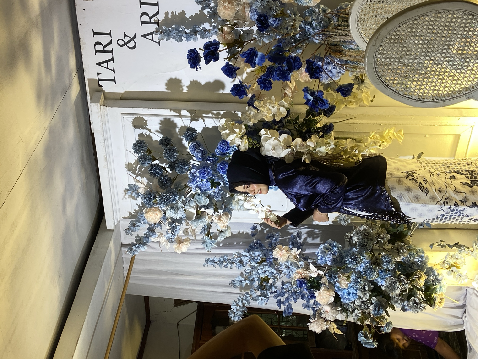 In Kembang Harum, Indonesia, a woman wearing a navy blue hijab and a matching satin jacket is posing for a photo in front of a decorative backdrop at what appears to be an event. The backdrop features an arrangement of artificial flowers in shades of blue, white, and peach, with some yellow floral accents hanging down. A white curtain hangs behind the floral arrangement. Text reading "TARI & ARI" is visible on the wall to the right of the backdrop. Two white chairs with woven seats are positioned to the right of the frame. To the left of the frame, a person in a purple shirt is partially visible, bending down. The lighting is warm and appears to be artificial, creating shadows on the wall. The ceiling shows exposed beams.