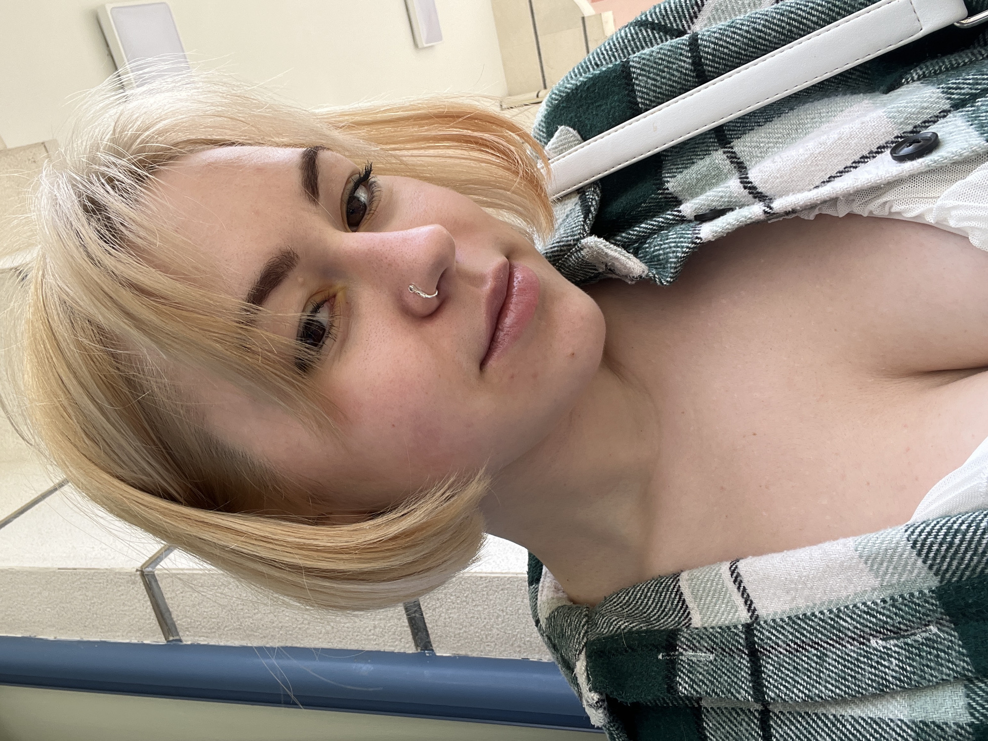 The image presents a young woman with blonde hair, fair skin, and prominent dark eyebrows, captured from a low, tilted angle. She is looking towards the camera with a neutral to slightly serene expression. A silver ring is visible as a piercing on her left nostril, and her lips appear glossy.
She is wearing an open green, white, and black plaid shirt or jacket, which partially reveals a hint of a white, possibly ruffled, garment underneath. A white strap, likely from a bag or purse, diagonally crosses her upper body.
The background suggests an indoor setting, with light-colored tiled surfaces visible, possibly on walls or flooring. A segment of a blue railing or banister can be seen towards the bottom of the frame, hinting at a hallway or stairwell environment. A rectangular light fixture is mounted on the wall in the upper portion of the background. The lighting in the image suggests daytime or a well-lit indoor space. The location context provided indicates the scene is in Budva, Montenegro. There is no visible text in the image.