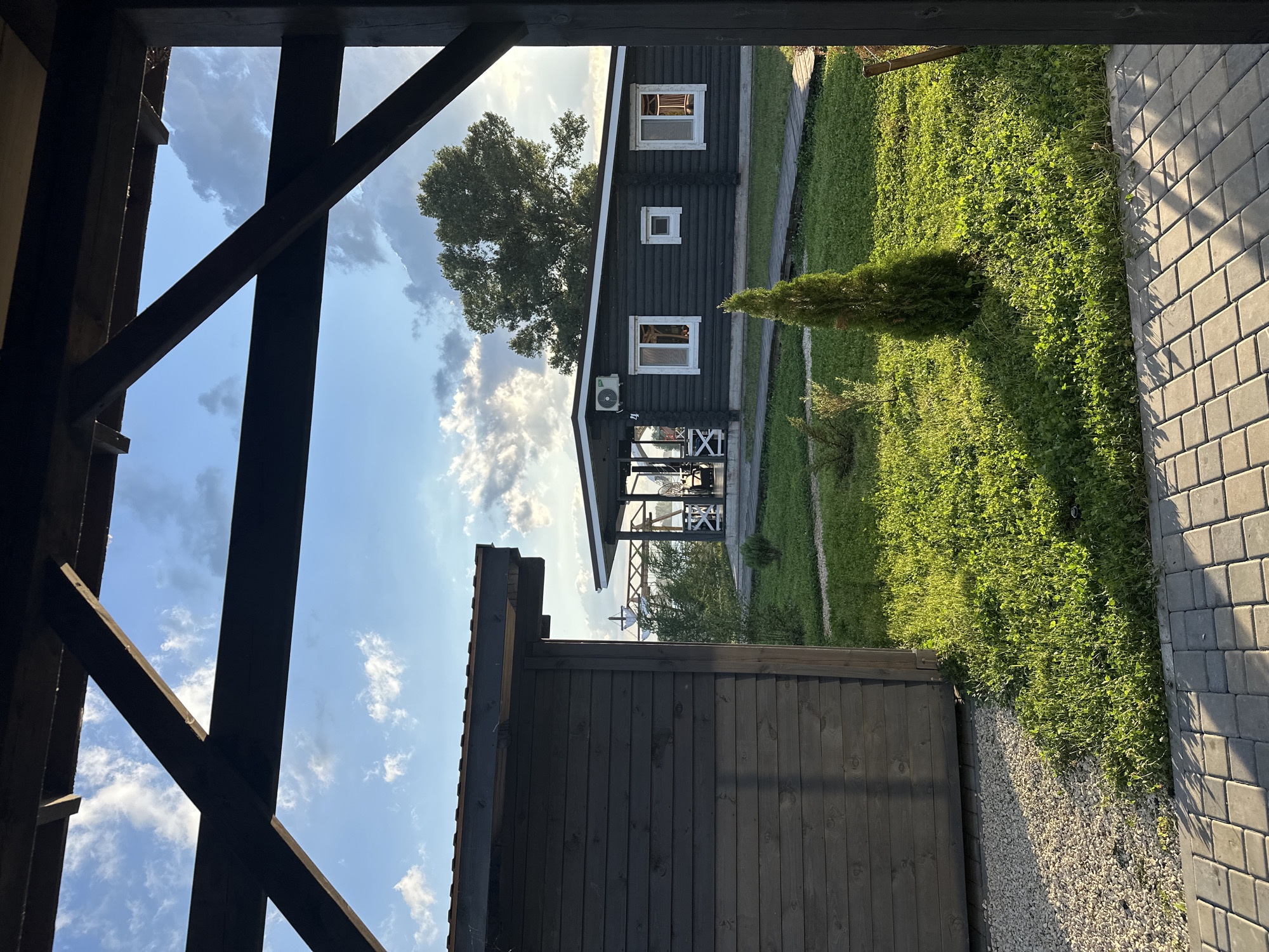 The image depicts a tranquil scene with two dark wooden structures set against a backdrop of a partly cloudy sky and lush green grass. In the foreground, a wooden pergola with dark beams frames the view. To the left, a dark wooden building with horizontal siding occupies a significant portion of the frame. To the right, a longer, low-slung dark wooden structure with a covered porch and white-trimmed windows is visible. The porch features white X-shaped railings. A small, conical evergreen tree stands in the well-maintained lawn between the buildings. A pathway made of light-colored pavers is visible in the lower foreground, leading towards the buildings. The scene suggests a resort or a residential area in a natural setting, possibly in Balakovo, Russia, given the context. There are no people visible in the image.