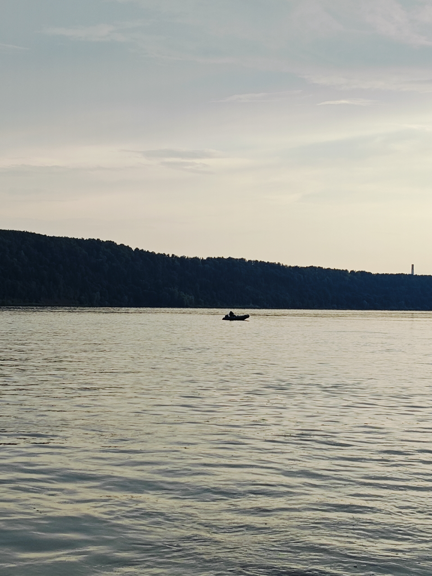 In Noviy, Russia, a tranquil scene unfolds on a wide body of water, likely a river, under a soft, overcast sky. The foreground is dominated by the calm surface of the water, reflecting the muted light with gentle ripples creating a textured pattern. Across the water, a densely forested hill forms a dark, undulating silhouette against the horizon, its trees creating a rich, deep green tapestry.
Towards the right side of the image, atop the hill, a solitary, tall, red and white striped tower stands out, piercing the sky. Closer to the center of the frame, a small, dark inflatable boat drifts on the water, carrying a single occupant silhouetted against the expanse. The overall atmosphere is one of quiet serenity, with the natural landscape and the solitary boat suggesting a moment of peaceful solitude.