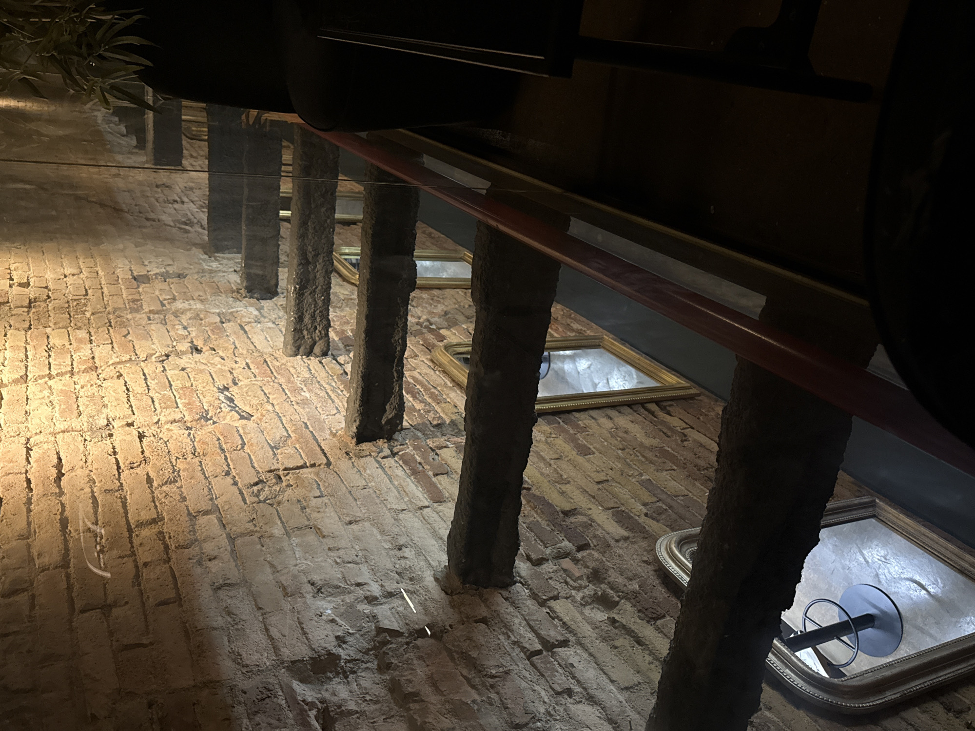 The image captures a close-up, high-angle view of an interior staircase with exposed brick walls. The staircase features rough, dark-colored steps that appear to be made of stone or concrete, supported by what seem to be thick wooden beams embedded in the brickwork. A red handrail runs along the side of the stairs, separated by a clear glass panel. Several ornate, gold-framed mirrors are visible on the brick wall below the stairs, reflecting the environment, including a bar stool. The lighting is dim, with a warm spotlight illuminating the brick wall and casting shadows, suggesting an evening or dimly lit indoor setting. The overall aesthetic is rustic and industrial, with a touch of vintage elegance from the mirrors. The location is not specified in the image itself.