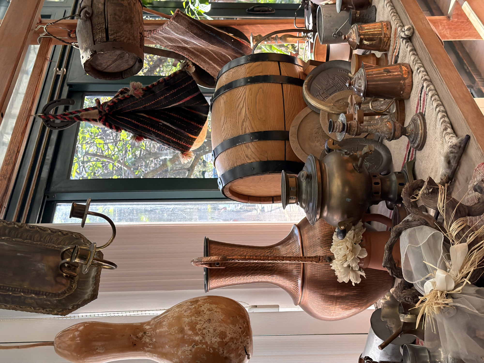 The image depicts a collection of antique objects displayed in an indoor setting. Objects include a wooden barrel, copper pots and jugs, a samovar, candle holders, and decorative items like dried gourds, fabrics, and floral arrangements. A window with a view of trees is visible in the background. Based on the types of objects and the overall aesthetic, it is plausible that the photo was taken at the Budva City Museum, which includes the Ethnographic Museum, in Budva, Montenegro.