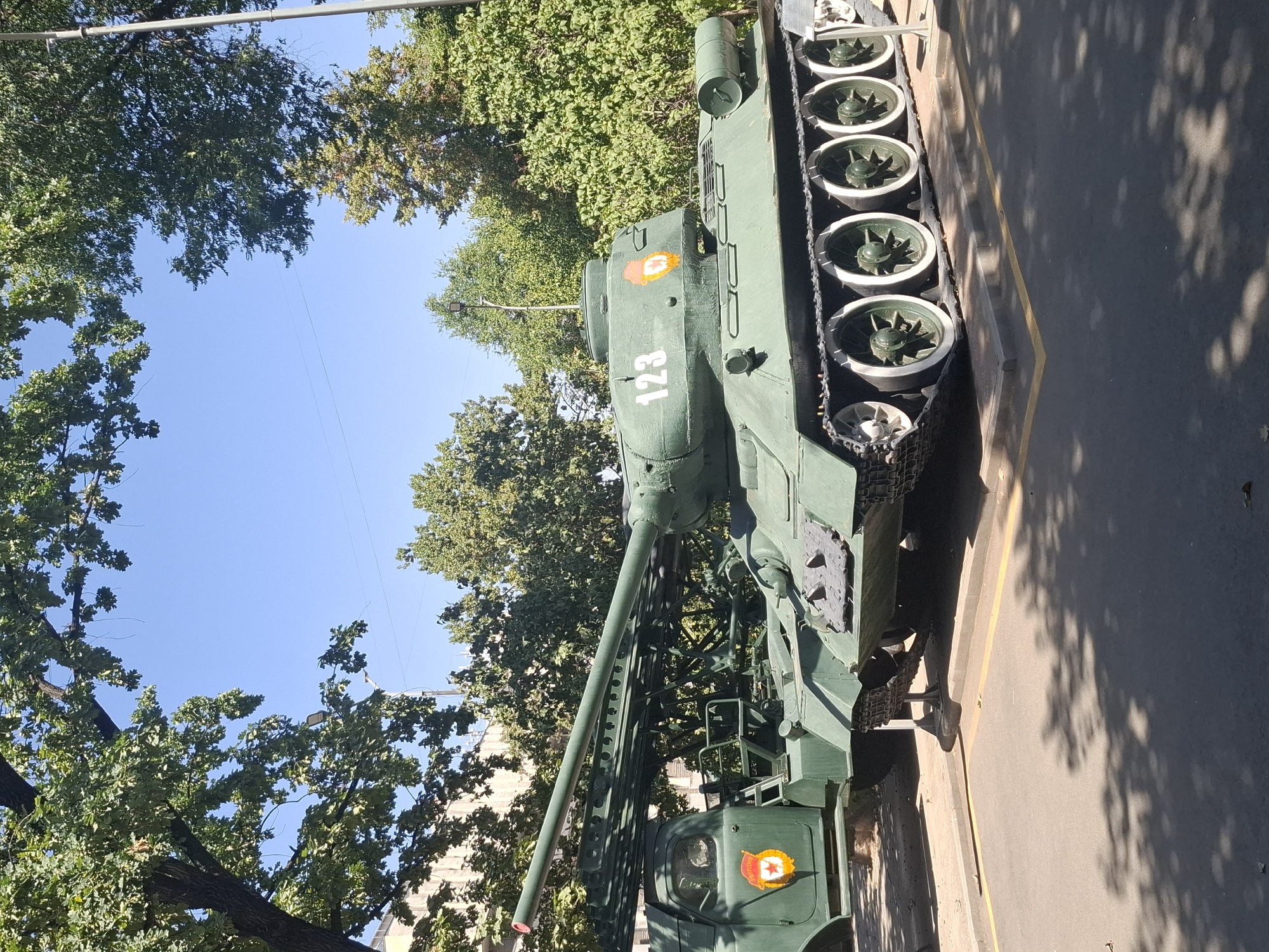 The image depicts a parked military vehicle, likely a Soviet-era T-34 tank, on a sunny day. The tank is painted in a matte green color and features white rims on its road wheels, a common modification. On the side of the turret, the number "123" is painted in white, and an emblem with a red star and orange rays is visible. Adjacent to the tank, a truck chassis with a rocket launcher system is also present, displaying a similar emblem on its door. The vehicles are positioned on a paved area with yellow painted lines, possibly in a display or memorial setting. Lush green trees with dense foliage form a backdrop against a clear blue sky. The lighting suggests it is mid-day. The environment appears to be an outdoor urban setting. Based on visual cues, specifically the emblems and vehicle type, this scene is consistent with the Memorial Complex of Glory (also known as the Eternal Flame) located in Panfilov Park in Almaty, Kazakhstan. This park is a prominent landmark in the city.