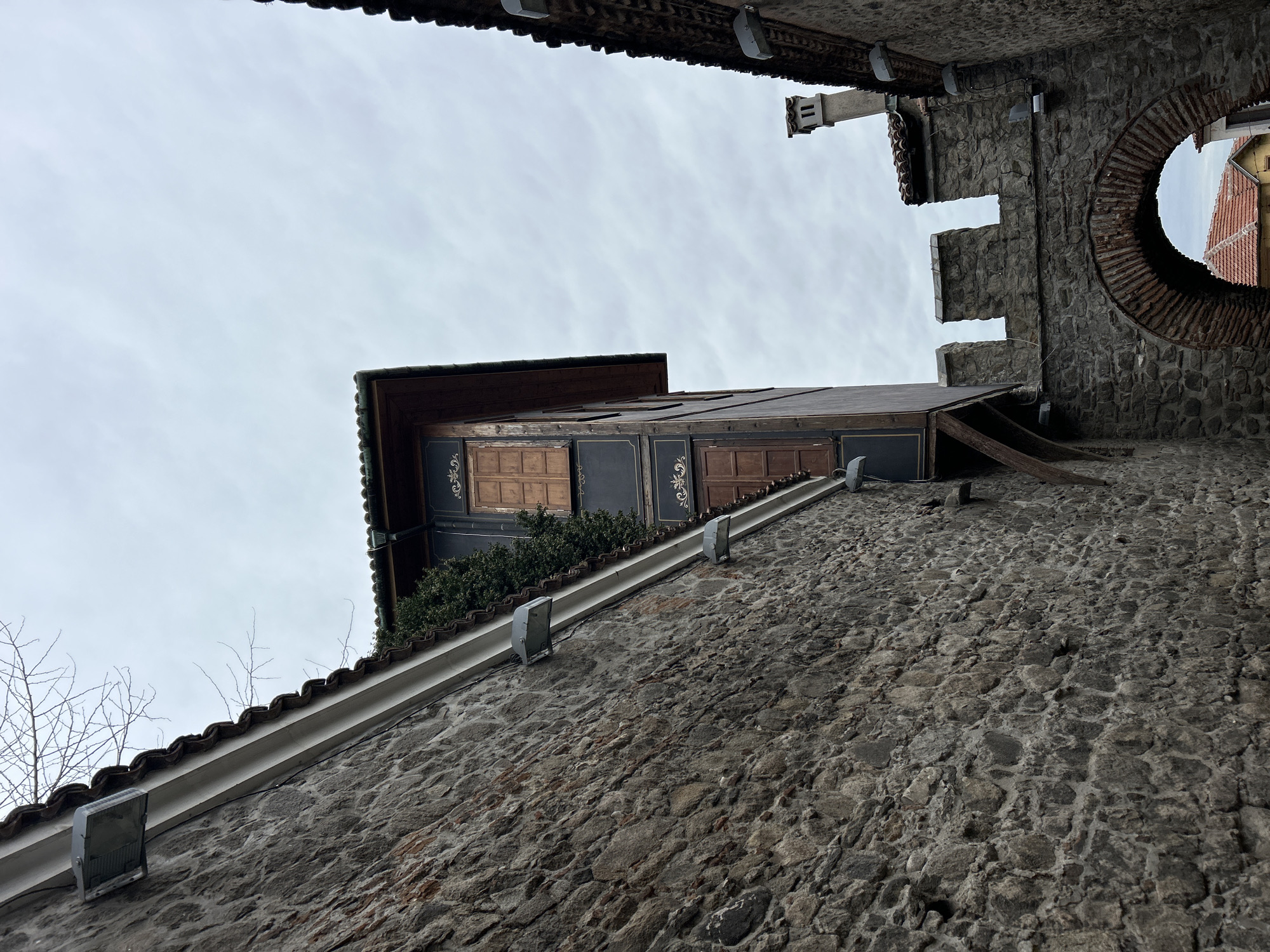 The image shows a view of buildings and architectural details in Plovdiv, Bulgaria, taken from a low angle looking upward. The primary subject is a stone wall with a white gutter and attached lighting fixtures. Above the wall, a traditional Bulgarian house with wooden shutters and decorative elements is visible. The house has a dark trim color with ornate gold detailing. A segment of a round brick archway is also seen, and the sky is overcast. The angle of the image is tilted.
