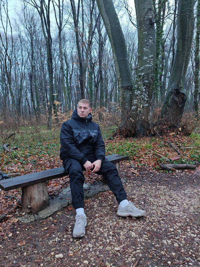 A single male individual is seated on a wooden bench in a forested area located within the city of Shumen, Bulgaria. The subject, appearing to be a young adult with a light complexion and short, closely cropped hair, is dressed in a black hooded puffer jacket featuring a dark, subtle camouflage pattern across the chest and shoulders. He wears black cargo-style trousers, white socks, and light grey athletic shoes. His hands are clasped loosely in front of him on his lap, and he is looking towards the viewer's left with a neutral expression.
The bench is constructed from dark-stained timber, supported by two robust, round log sections. The ground surface directly beneath and surrounding the bench is a mixture of small gravel, soil, and fallen brown deciduous leaves, suggesting an autumn or winter season. Sparse green vegetation is visible amidst the leaf litter.
The background consists of a dense arrangement of bare deciduous trees, devoid of foliage, confirming the seasonal indication. A large tree with a distinctive multi-trunk formation (appearing as three substantial trunks) stands prominently to the right of the seated individual, its bark displaying lighter patches. The sky is overcast, providing diffuse, cool lighting to the scene. No other individuals or significant objects are present.