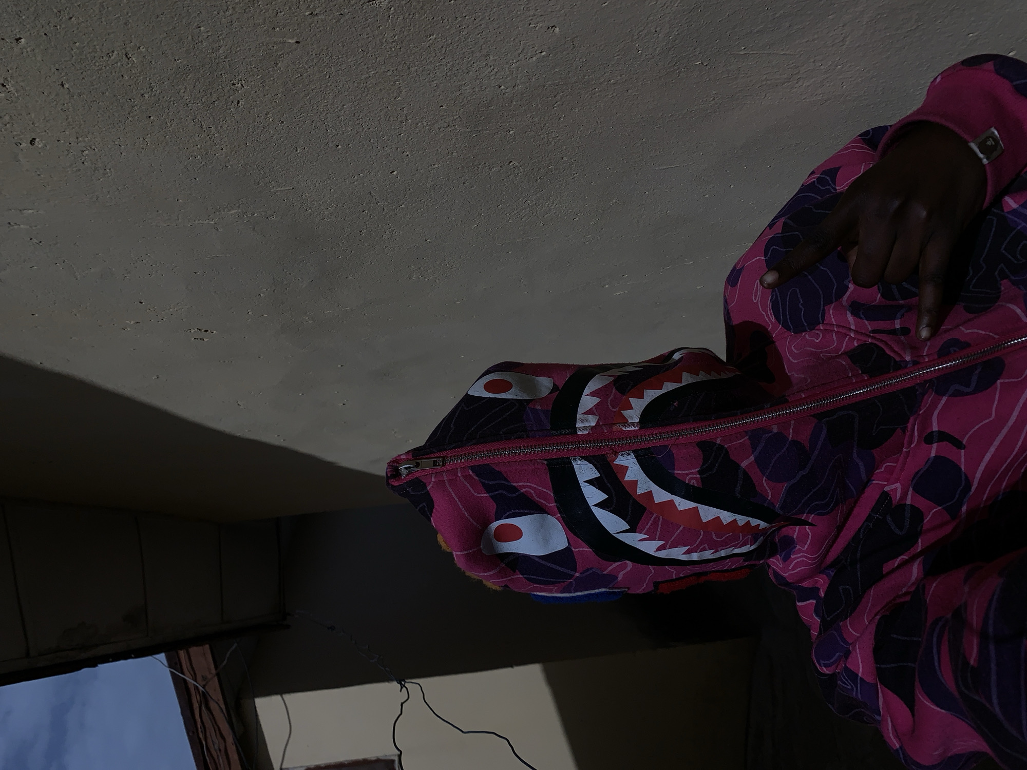 The image depicts a person wearing a pink and purple camouflage patterned hoodie with a shark design on the hood. The person's hand is visible, making a hand gesture. The background includes a ceiling and part of a window frame. The location is not identifiable as Rafin Par, Nigeria, without additional context or visual cues.
