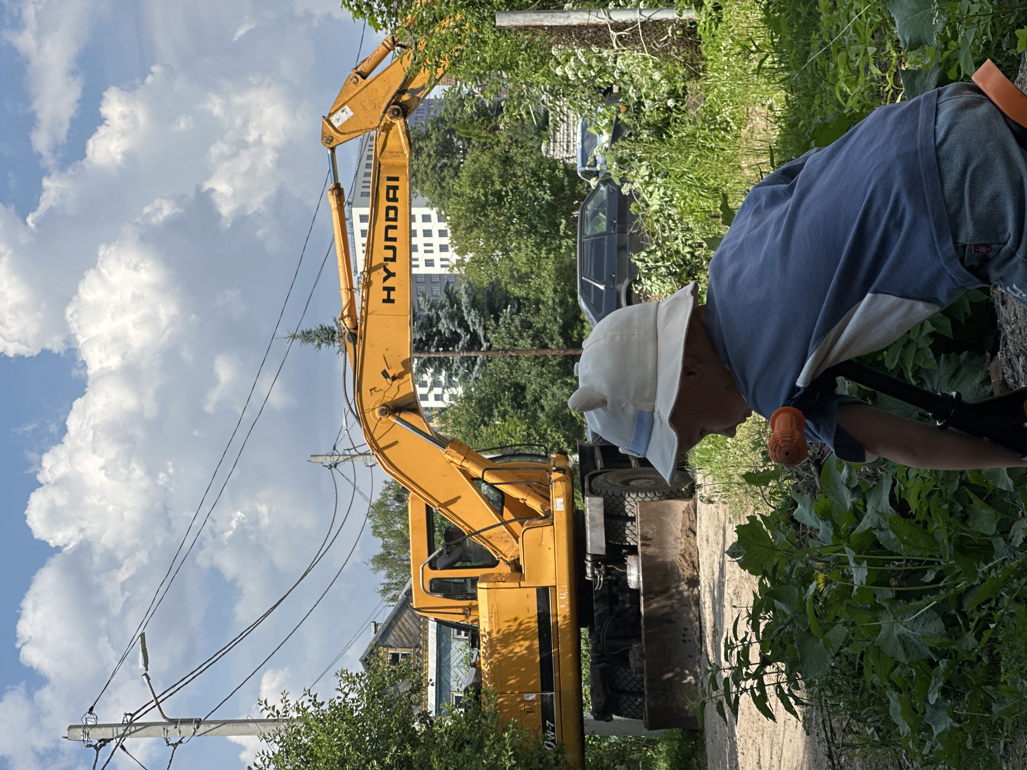 A young child wearing a white bucket hat with light blue trim and a blue t-shirt is partially visible in the foreground, seen from behind, looking towards a large yellow Hyundai excavator parked on a dirt path. The excavator is the main subject, its arm extended and a blade in front. Behind the excavator, a multi-story building with white windows and a lighter colored facade is visible, alongside several trees. Other elements in the background include power lines suspended from a utility pole, a dark-colored sedan, and a light-colored car. The scene appears to be an outdoor area with a mix of natural vegetation and construction or maintenance equipment, possibly in the city of Nizhny Novgorod, Russia, given the context. The sky above is partly cloudy with large white cumulus clouds.
