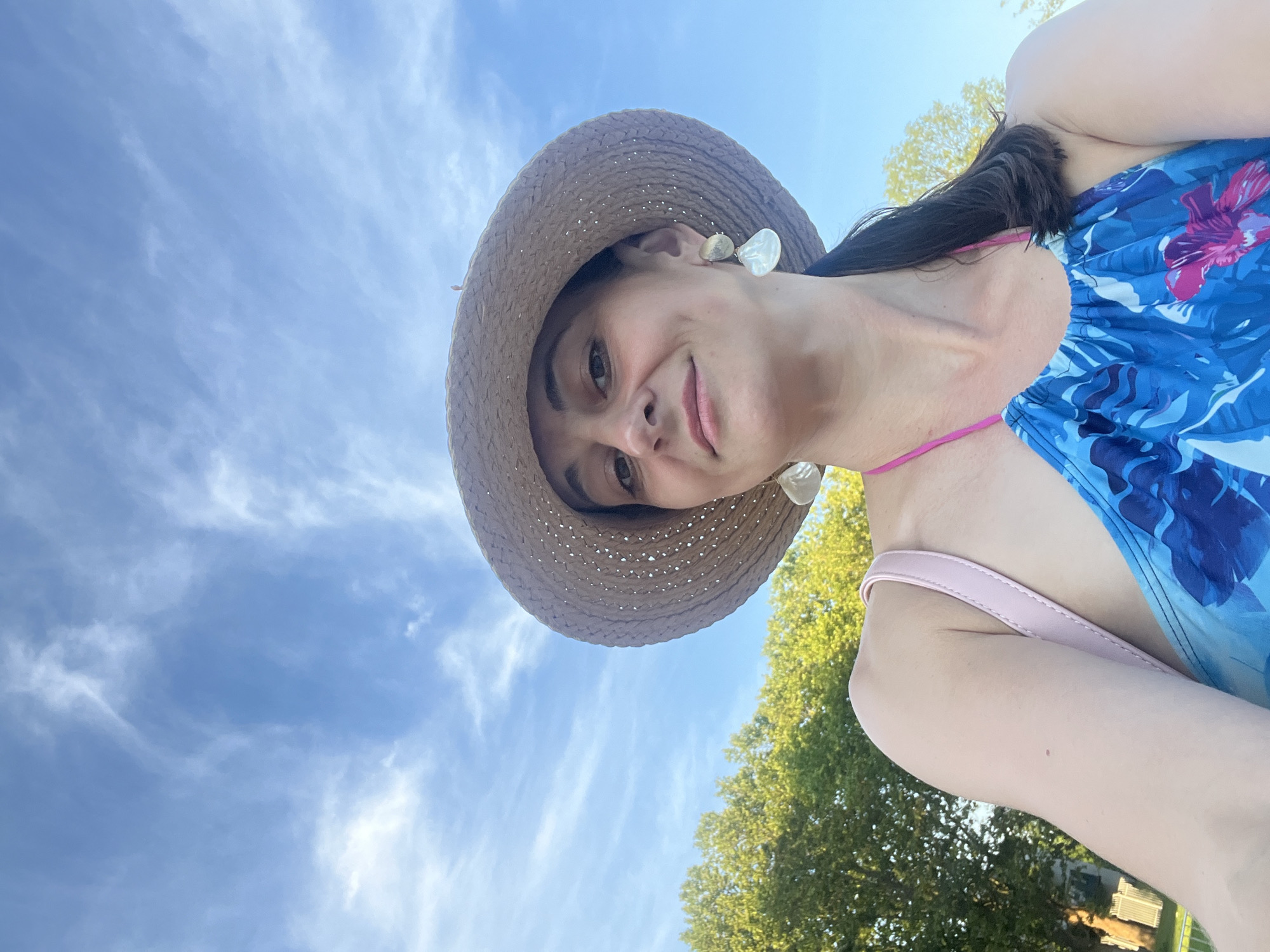 A woman is taking a selfie outdoors. She is wearing a straw hat, a blue floral swimsuit with a pink string, and earrings made of shell. Behind her, the sky is blue with wispy white clouds, and green trees are visible. The image is taken from a low angle, looking up at the woman and the sky. The woman has dark hair pulled back into a ponytail and is smiling at the camera.