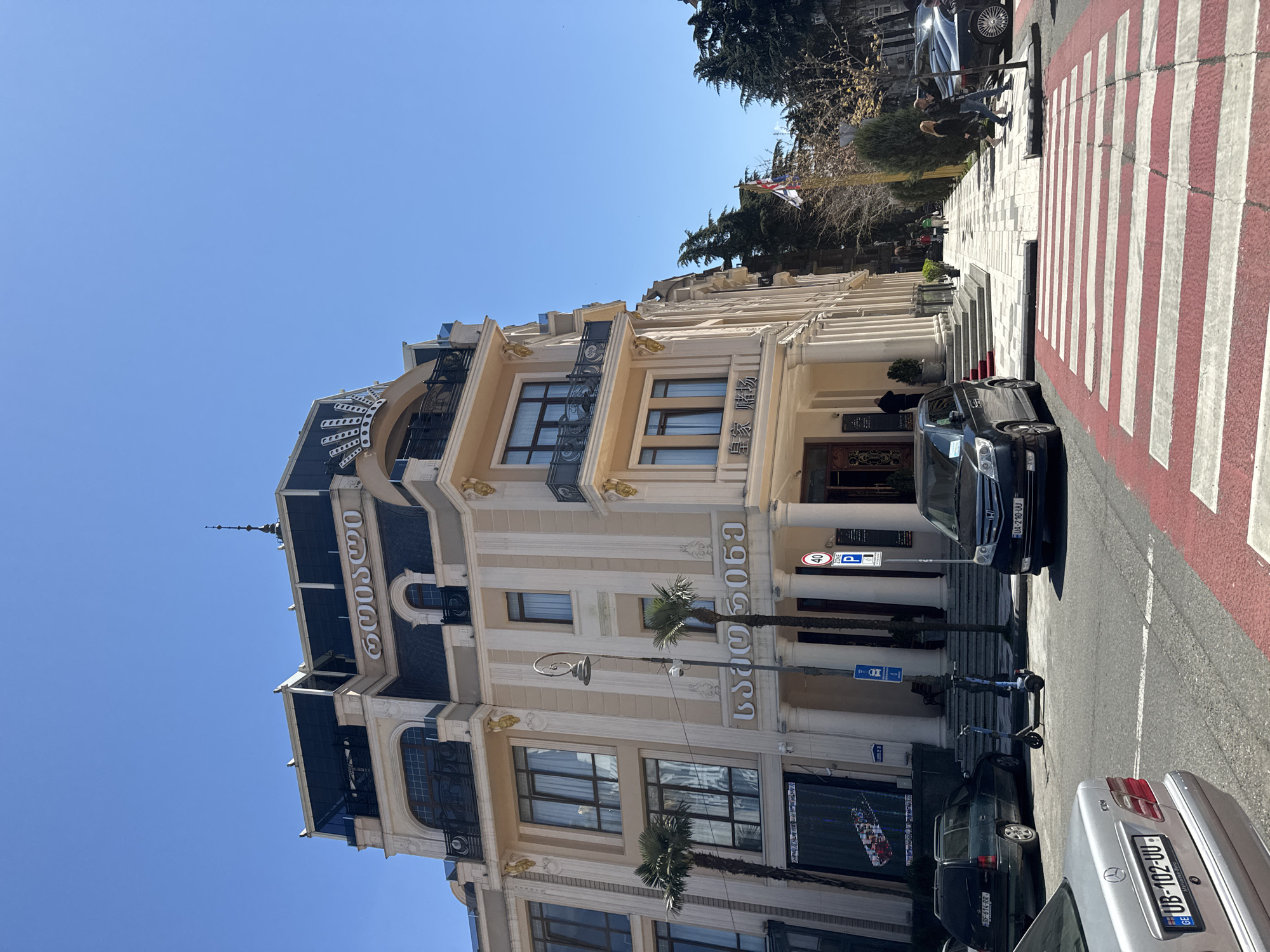 The image shows a building with elaborate architectural details in what appears to be a city setting. The building has a beige facade with ornate gold detailing and what appears to be dark blue accents or roofing. There are Georgian and Chinese words displayed vertically on the building. Palm trees are planted near the base of the building, adding a touch of tropical urban landscape.
Several vehicles are parked along the street, including a silver Mercedes Benz with license plate "UB-102-UU," and a black car with license plate "UA-210-UU." Scooters and people are on the sidewalk. The street has a red and white striped crosswalk. A street sign with "40" in a circle and parking symbols indicates a restricted zone.