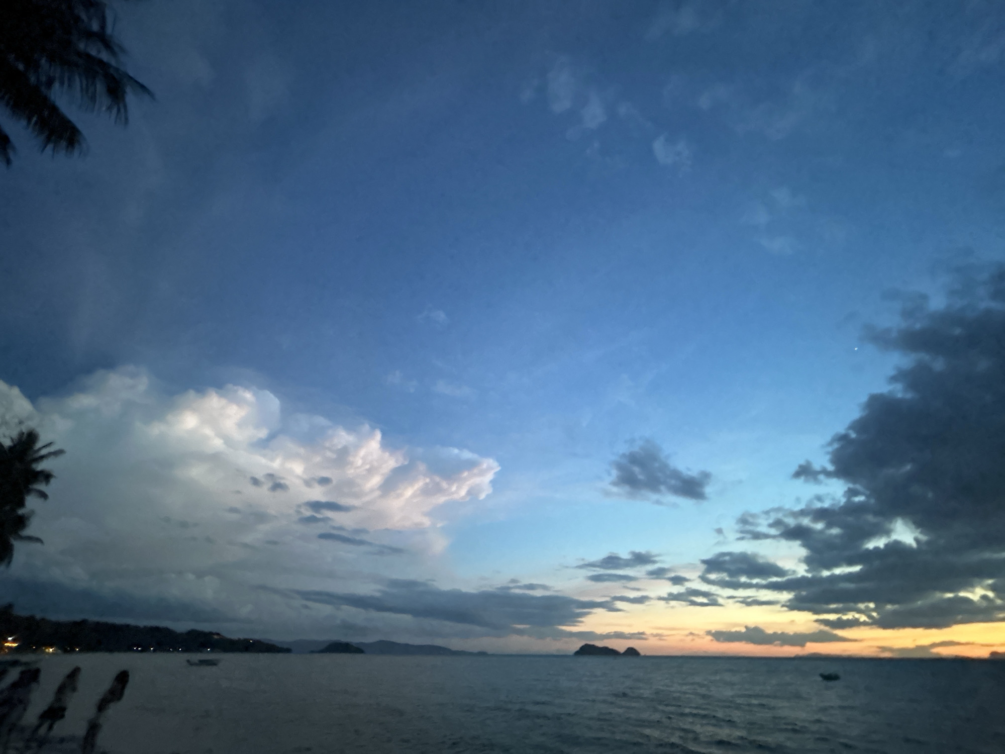 A wide-angle, low-light, medium-shot captures a tranquil beach scene in Ko Pha-ngan District, Thailand, during twilight. The sky transitions from a deep twilight blue at the top to a soft gradient of orange and yellow near the horizon. Large, dramatic clouds dominate the left side of the frame, with some illuminated by the fading light, while darker clouds gather on the right. The sea is calm, with gentle ripples on its surface. In the distance, silhouetted islands rise from the water, and a faint line of lights suggests a coastal settlement. A solitary boat is visible on the water. The foreground is a beach, with the dark silhouettes of palm fronds from a tree on the far left partially obscuring the sky. A few indistinct figures, also silhouetted, are present on the beach. The overall mood is peaceful and serene.