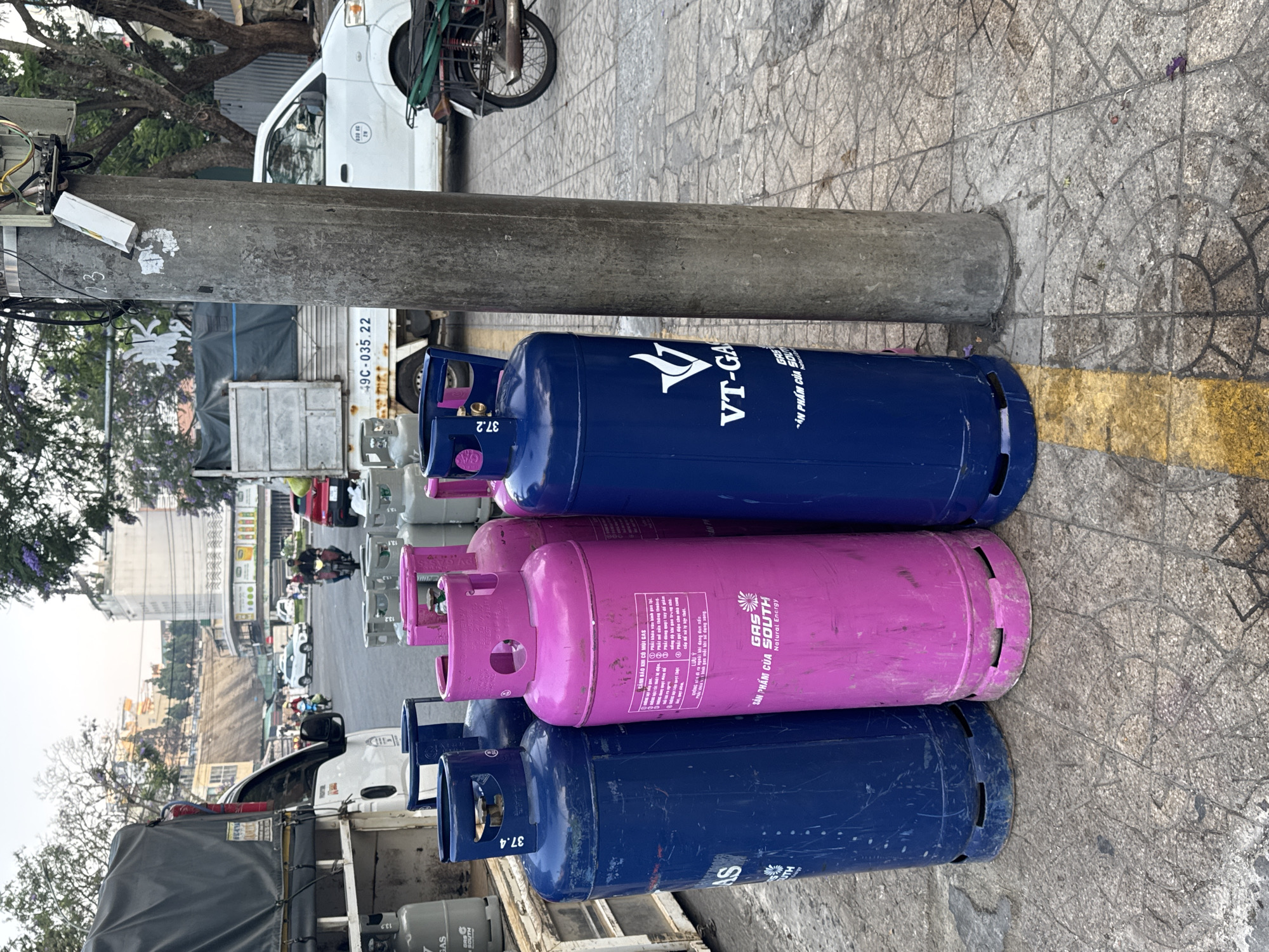 The scene shows three gas cylinders stacked on a sidewalk next to a concrete pole in what appears to be Da Lat, Vietnam. The cylinders are blue and pink, with one blue cylinder on top, a pink one in the middle, and another blue cylinder at the bottom. The cylinders have writing on them in Vietnamese and English, including brand names like "VT-GA" and "GAS SOUTH." A truck is partially visible in the background, with other gas cylinders loaded onto it. The sidewalk has a patterned tile surface and a faded yellow line. Other vehicles, buildings, and trees are visible in the background, suggesting a busy urban environment. The license plate number 49C-035.22 is also visible on a vehicle.