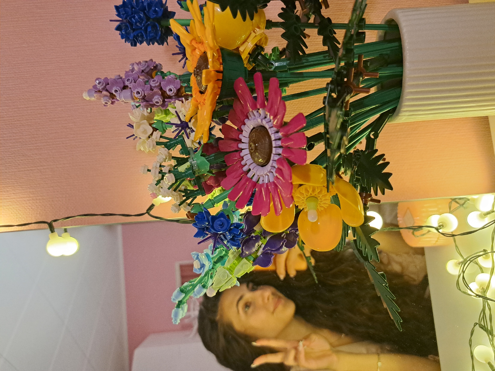 An image, rotated 90 degrees counter-clockwise, depicts a vibrant bouquet of artificial flowers, likely made from building blocks like Lego, arranged in a white, ribbed vase. The flowers are multi-colored, featuring bright pink, yellow, blue, purple, and white blossoms with green stems and leaves.
Below the vase and flowers, a mirror reflects a young woman with long, dark, wavy hair. She is looking upwards, seemingly at the flowers or the camera, and is making a peace sign with her right hand, which has a ring on a finger and painted nails. Her expression is a soft smile.
The setting appears to be indoors, illuminated by warm string lights that are visible both directly and in the mirror's reflection. The background behind the flowers is a plain light pink or peach wall, while the reflection shows parts of a room, including what might be furniture or a bed. The string lights add a cozy, evening ambiance to the scene. No text is visible in the image.