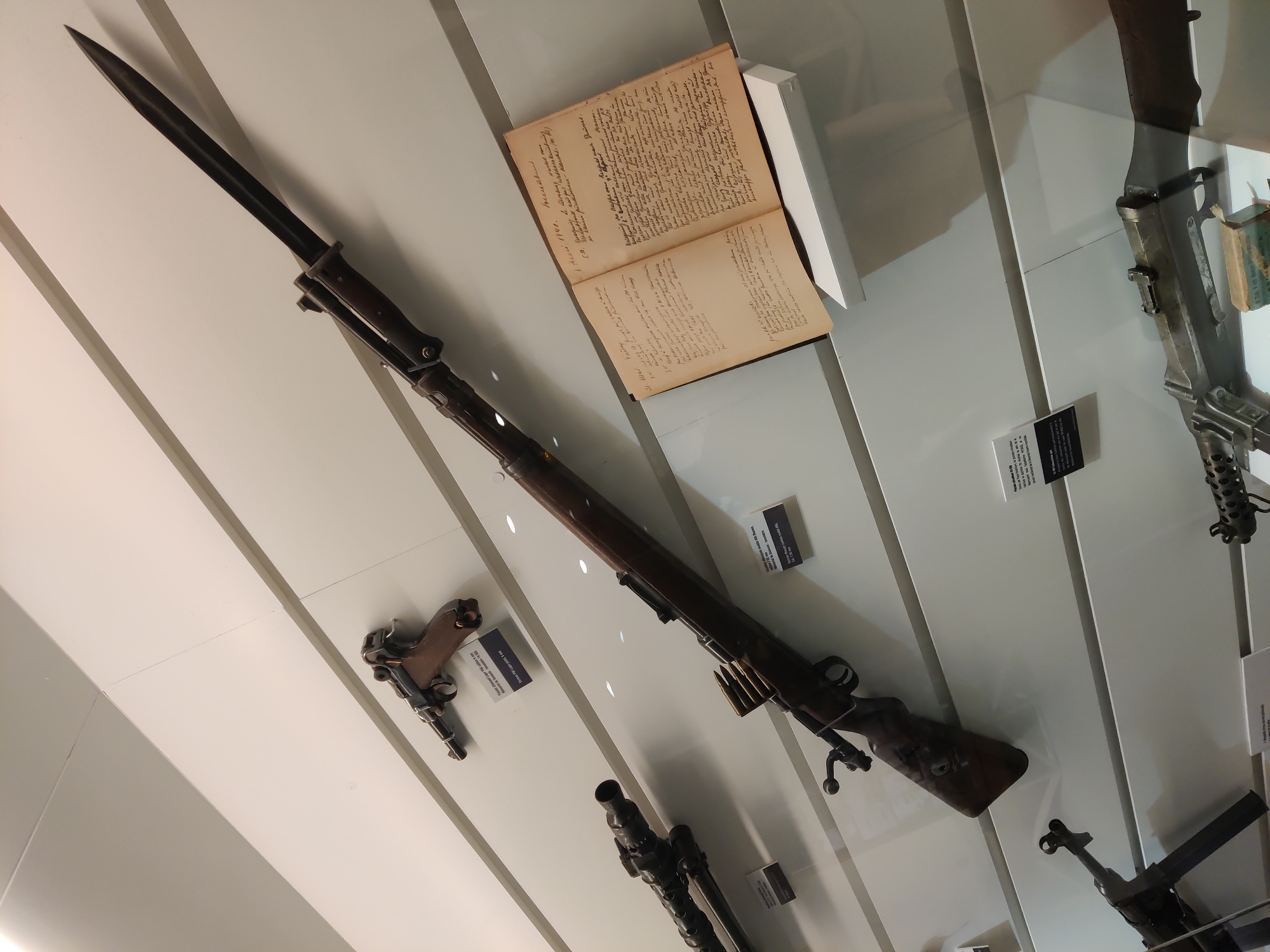 The image displays a collection of historical firearms and a notebook, likely exhibited in a museum. The primary focus is on a rifle with an attached bayonet, positioned diagonally across the frame. To the left and slightly above it, a pistol is mounted. Below the rifle, another firearm, possibly a machine gun, is partially visible. To the right of the rifle, an open notebook with handwritten text is displayed on a white stand. The background consists of white, angled panels, suggestive of a museum display. The overall lighting is bright, illuminating the objects. The location is identified as Dunkirk, France, although no specific landmarks or context within Dunkirk are visually apparent. There are no people or discernible actions taking place. The scene is static and observational.