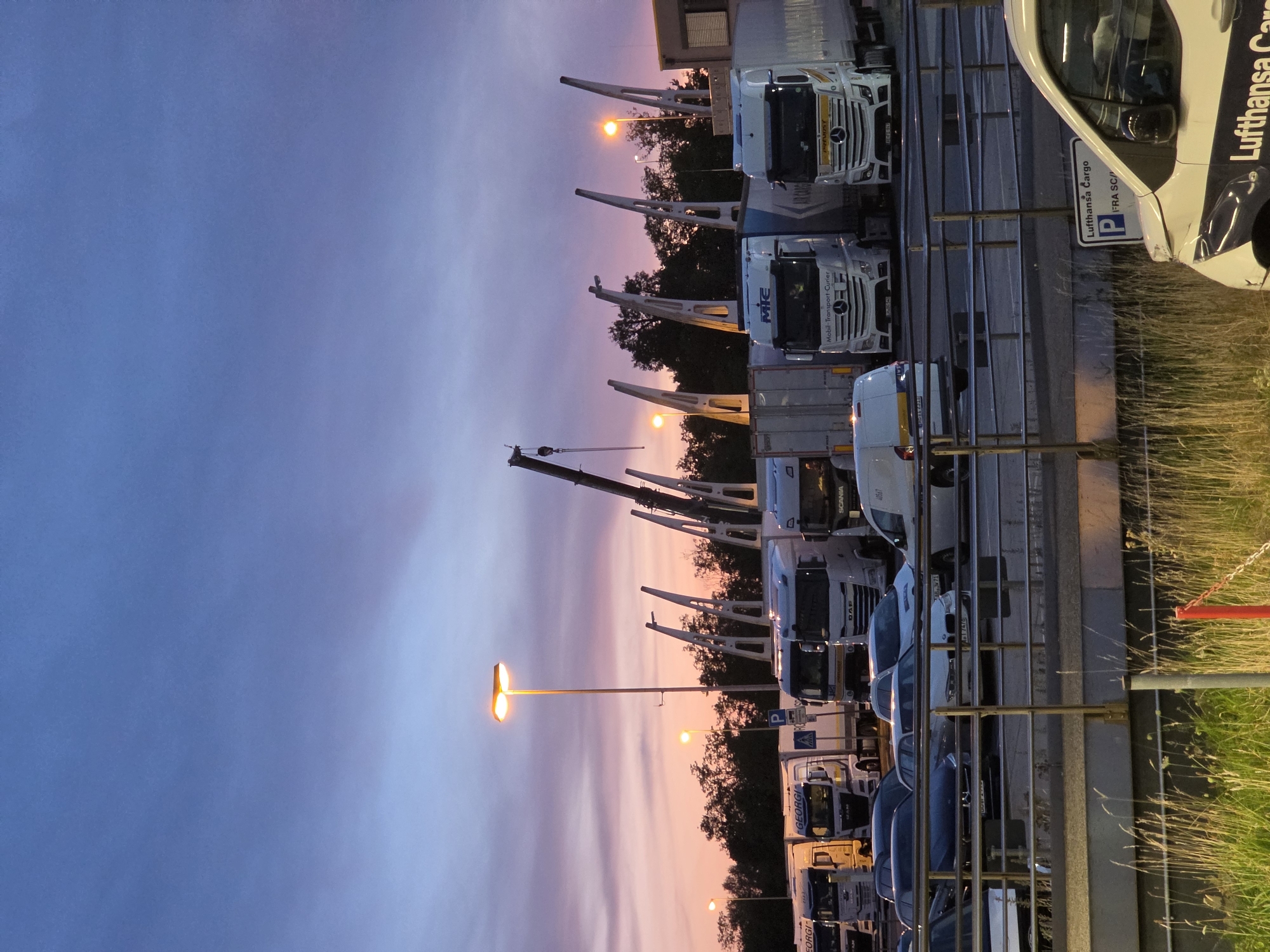 The image depicts a fleet of parked trucks and vehicles at what appears to be a cargo terminal or logistics hub at dusk. The sky displays a gradient of blues and purples with hints of pink, suggesting either sunrise or sunset. Several large transport trucks, some with trailers, are lined up in a lot. Among them are vehicles from brands like Mercedes-Benz, DAF, and MAN, some featuring company logos such as "GEORGI" and "MIC - Mobil-Transport-Curier". A white van with "Lufthansa Cargo" branding is partially visible in the foreground on the right. In the background, there are trees and a building. Cranes are also visible, implying a loading or unloading area. The scene is set on what appears to be an airport apron, consistent with the Lufthansa Cargo branding, suggesting a location at Frankfurt Airport, Germany. There are no people clearly visible in the image. The environment is illuminated by streetlights and the natural light of the twilight sky.