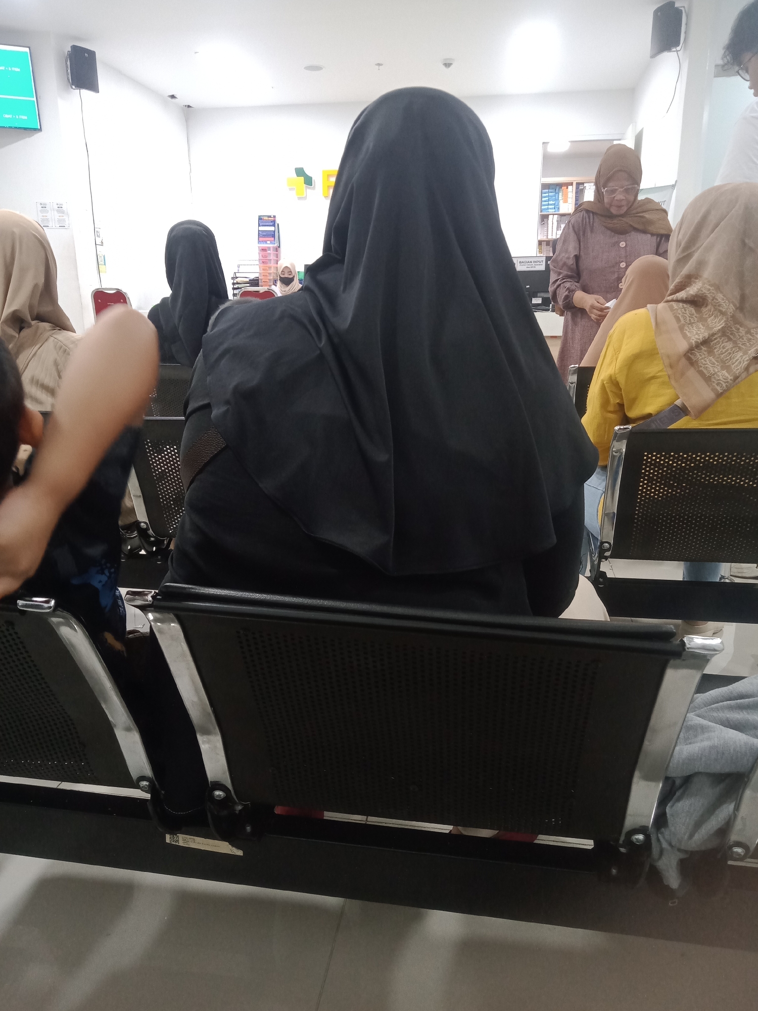 The image depicts a waiting area, likely at a medical facility, with several individuals seated and one standing. The primary subject in the foreground is a person wearing a black hijab, facing away from the camera. To their left, another person in a light brown hijab is seated. To the right of the person in the black hijab, another individual in a yellow top and beige hijab is visible.
Further back, a woman in a brown headscarf and glasses stands and appears to be interacting with someone off-camera or attending to a task. A shelf unit filled with items, possibly medical supplies or records, is visible in the background, along with signage that suggests an administrative area. The environment features white walls, a tiled floor, and rows of black chairs with metal frames, indicative of a waiting room setting. The lighting appears to be artificial.
Based on contextual clues such as the signage with Indonesian text (e.g., "BAGIAN INPUT"), the presence of individuals wearing hijabs, and the overall aesthetic, this scene is likely located in Sumedang, Indonesia, within a public institution such as a clinic or hospital.