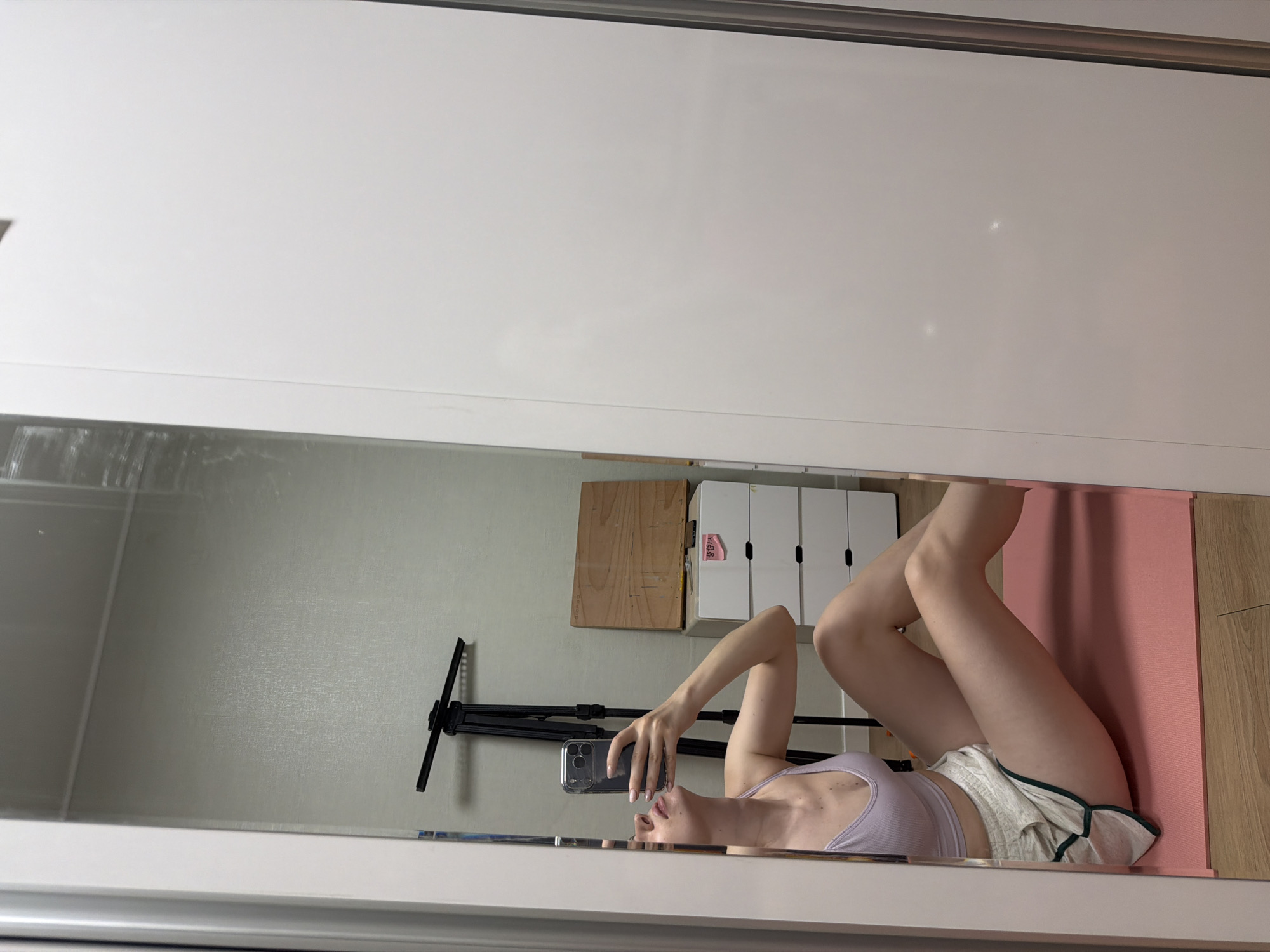 A young woman is taking a mirror selfie in her room, which appears to be in an apartment in Dongducheon-si, South Korea. She is sitting on a pink yoga mat with her legs bent and her body angled towards the mirror. She is wearing a light purple sports bra and white shorts with green trim. The mirror reflects her upper body and face as she looks up towards the phone she is holding to take the picture. In the background, visible in the mirror, is a tripod, a wooden board leaning against a chest of drawers, and a wall with textured wallpaper. The room's flooring is wooden.