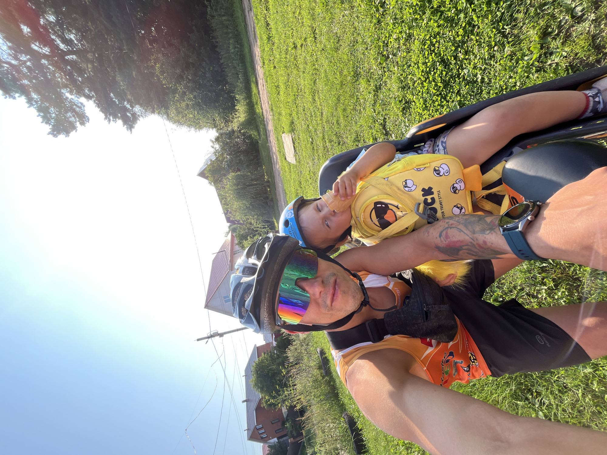 In Chaikovsky, Russia, on a sunny day with a clear blue sky and bright sun, a man wearing a cycling helmet, reflective sunglasses, and an orange tank top is taking a selfie. He is seated on a bicycle, and behind him, in a child seat attached to the bicycle, is a young child. The child is wearing a blue helmet and a yellow backpack with a duck print, and is eating an ice cream cone. They are in a grassy area, with houses and trees visible in the background. The man's arm is extended to take the photo, and a tattoo is visible on his arm. The overall scene depicts a father and child enjoying an outdoor activity, likely a bike ride, on a warm day in a residential area.