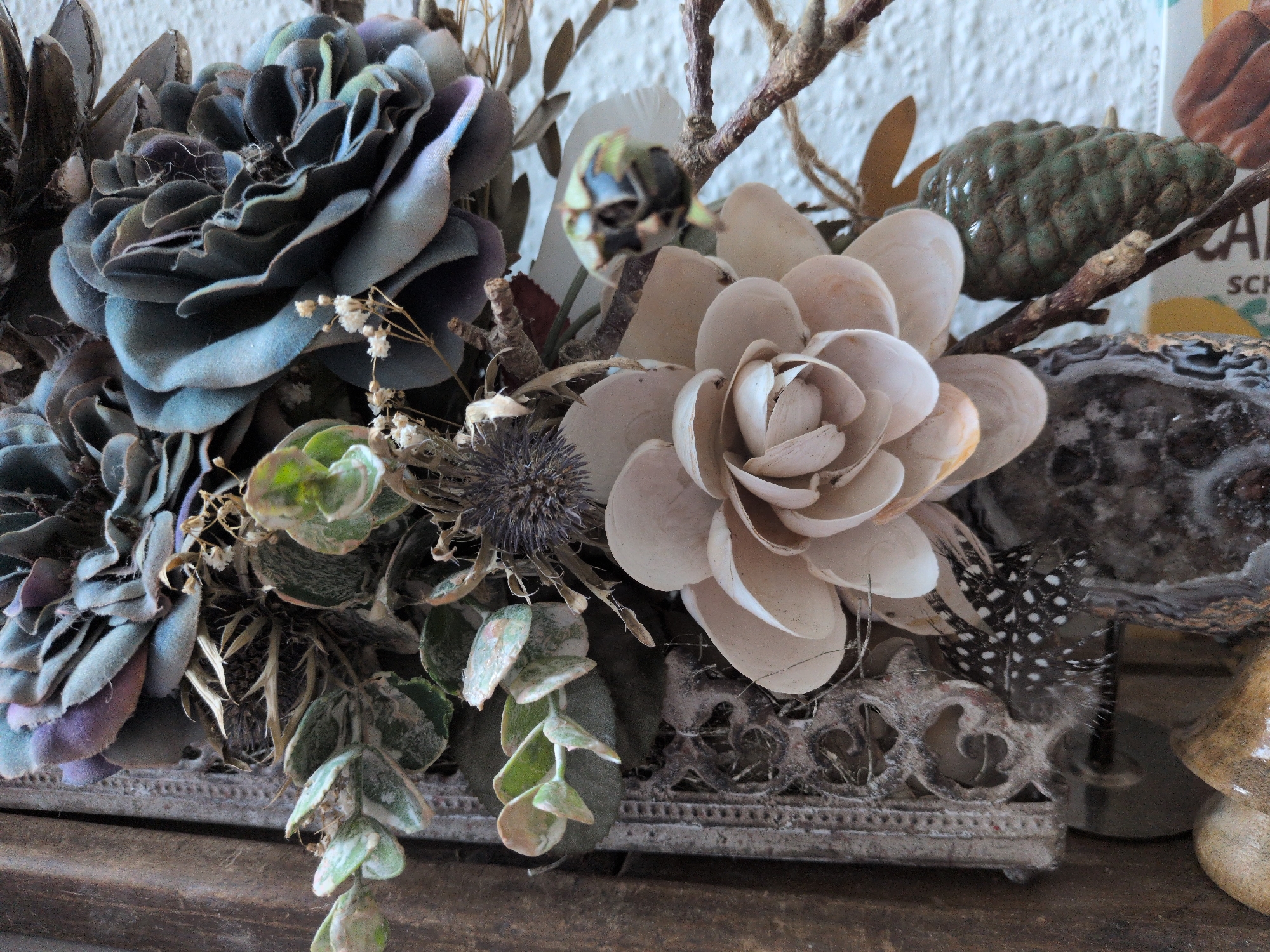 The image depicts a close-up still life arrangement of artificial flowers and natural elements, possibly as a decorative centerpiece. The dominant subjects are several large, multi-petaled artificial flowers in muted tones of dark blue, grey, and beige, with some exhibiting a velvety texture. Interspersed among the flowers are smaller, dried sprigs with tiny white blossoms, dried seed pods, and a few green succulent-like leaves. A dark, spiky dried flower head is also visible.
Further into the arrangement, a green, textured object resembling a small pinecone or a ceramic ornament is present. To the right, a spotted feather, likely from a guinea fowl, adds a textural contrast. The arrangement sits within an ornate, grey, rectangular container with decorative cutouts. In the background, a white textured wall is partially visible, along with a section of what appears to be a cardboard or paper surface with faded text, possibly from packaging. The overall impression is one of rustic and whimsical decor. There are no people, actions, or interactions depicted in the scene. The image is a still life and does not provide any information about the city of Brandenburg an der Havel, Germany, or its locations.