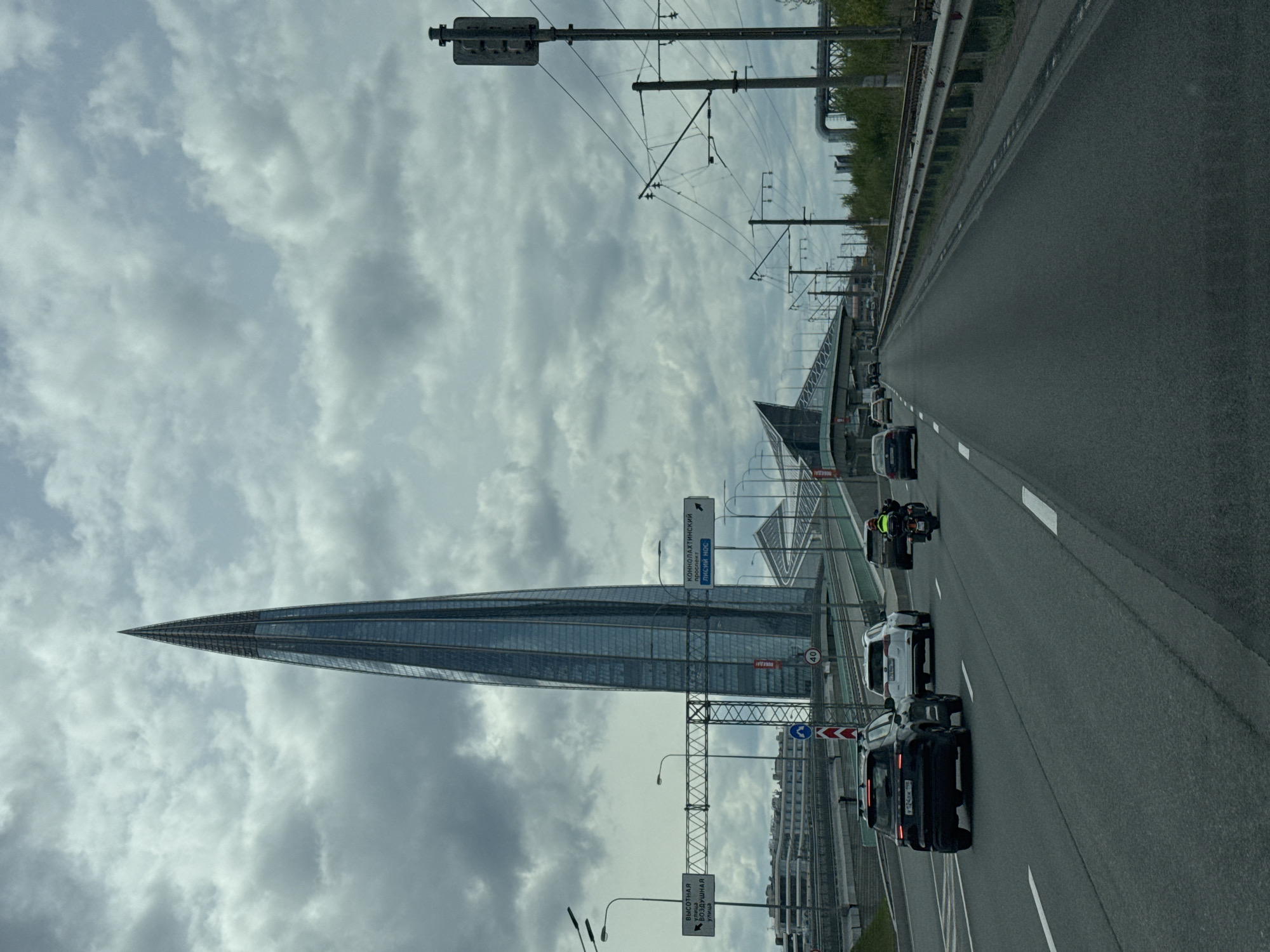 The image shows a highway in Saint Petersburg, Russia, with the Lakhta Center skyscraper prominently visible. The skyscraper is a modern glass structure that tapers to a point. The sky is overcast with gray clouds. Several vehicles, including cars and a motorcycle, are on the highway. Overhead electrical wires are present, likely for public transportation. Road signs are visible along the side of the road. The environment appears urban, with buildings lining the highway.