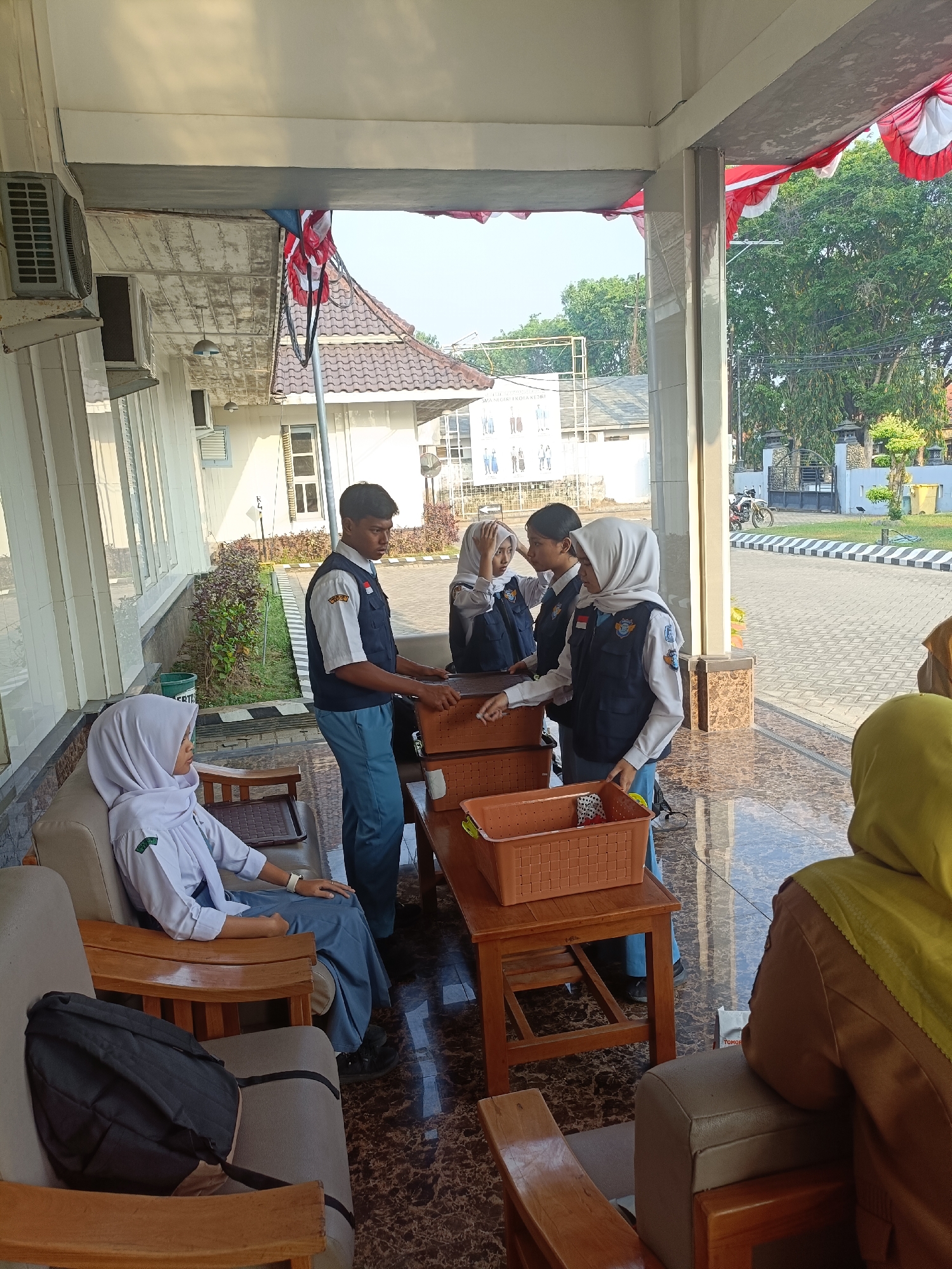 Four teenagers, likely students, are gathered on an outdoor patio area. Three of them, wearing uniforms consisting of white shirts, dark vests, and blue pants or skirts, are interacting around a wooden table. One male student stands, appearing to hand something to one of the female students, who is wearing a hijab. Another female student, also in uniform and wearing a hijab, is seated on a couch to the left, looking away from the group. A fourth individual, wearing a yellow hijab and brown top, is seated on a couch to the right, facing away from the camera. The environment appears to be part of a building with a covered walkway, featuring architectural details such as pillars and air conditioning units. In the background, trees and a street are visible, suggesting an urban or semi-urban setting. The presence of Indonesian flags suggests a national or patriotic context. Based on the building style and surrounding elements, this scene is likely located in Kediri City, Indonesia, potentially at an educational institution or public building.