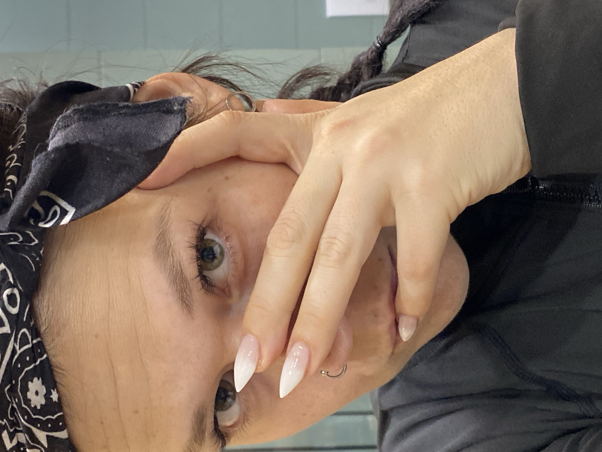 A close-up, rotated image shows a person with a black and white paisley bandana covering part of their forehead and hair. Their skin is fair with visible freckles. The person has light-brown eyes. Their right hand covers part of their face, with sharp, white-tipped fingernails. A silver hoop earring is visible in their ear. The person is wearing a black garment. The background is a blurred light green. There is no discernible information to indicate that the image was taken in Chaikovsky, Russia.
