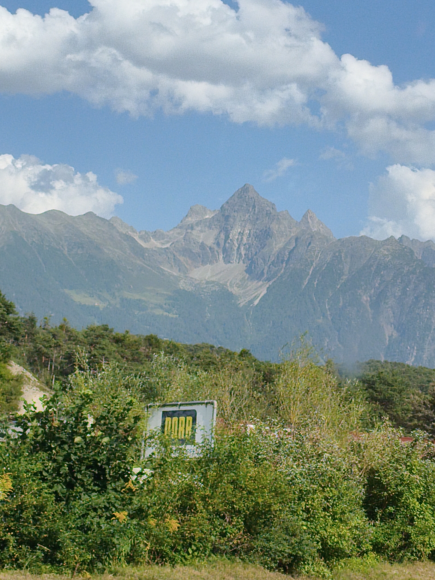 The image displays a mountainous landscape with lush green foliage in the foreground. A sign with the word "ROPA" in yellow block letters against a white background is partially obscured by bushes. The mountains, with their rocky peaks, are covered in trees and appear to be in Austria. The sky is blue with scattered white clouds.