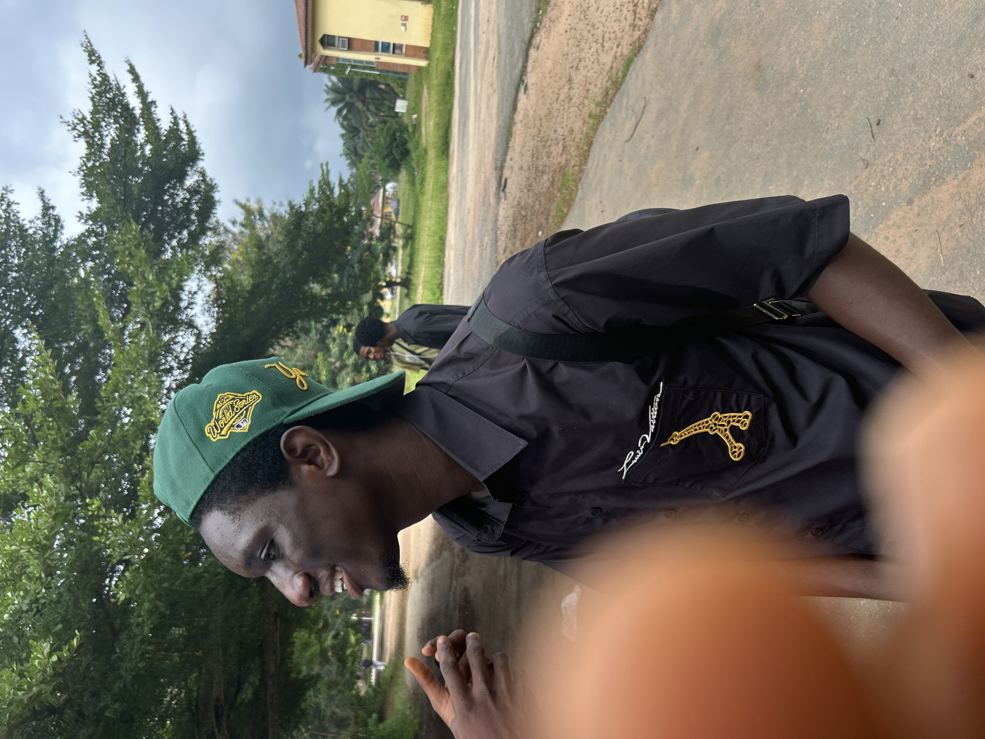 This is a portrait of a young Black man wearing a green baseball cap and a black short-sleeved shirt. He is outdoors, with lush green trees and a building in the background. The sky is overcast, suggesting a cloudy day. The man is looking down and to his left with a slight smile, revealing his teeth. He has a goatee. The shirt has a gold embroidered logo on the left chest, which appears to be a stylized Eiffel Tower with script text below it that reads "Louis Vuitton". Another person is visible in the background, also wearing a black shirt. The ground is paved with concrete and dirt. The overall impression is casual and relaxed. The location is Ihiagwa, Nigeria.