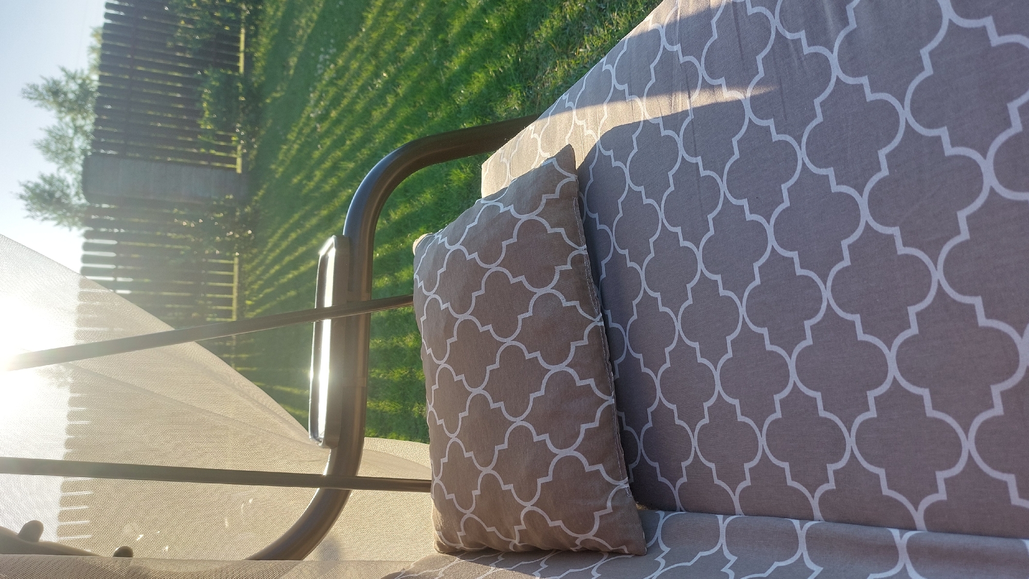 A close-up shot shows a cushion on a swing seat with a brown and white quatrefoil pattern. The cushion is placed on a larger seat cushion with the same pattern. A white mosquito net hangs over the seat, creating a soft, diffused light. In the background, a wooden fence encloses a green lawn, with trees visible behind the fence. The sun shines brightly, casting long shadows across the grass. The location is Masty, Belarus.