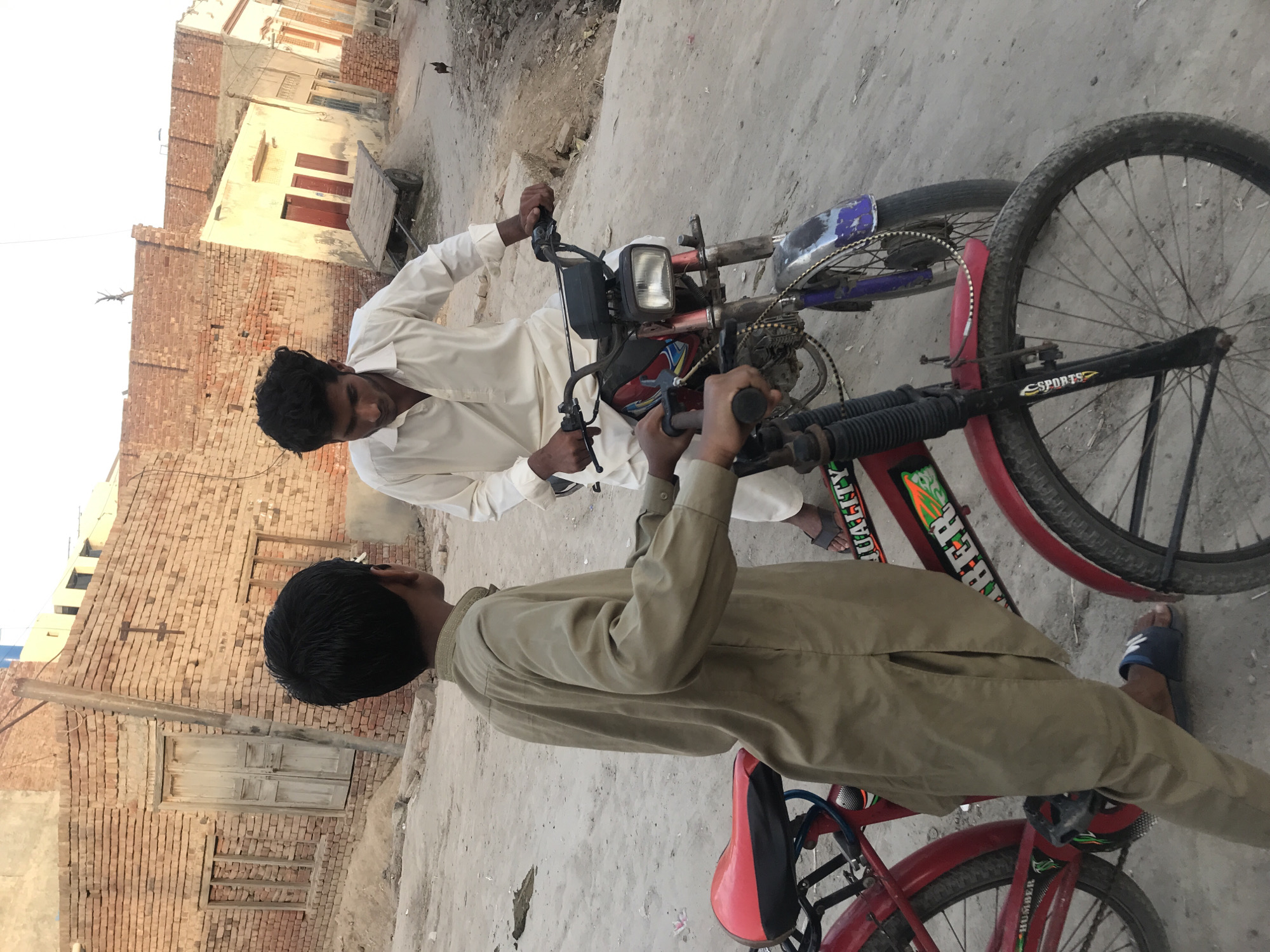 Two males are captured in a daytime scene on a dusty, unpaved street, likely in a South Asian locality. The setting features a row of simple, mostly brick buildings in the background, some with light-colored plaster finishes and wooden doors and windows. A utility pole stands near one of the structures, and a small, wooden pushcart is visible further down the street. The sky appears clear, indicating sunny weather.
On the left, a person wearing a white traditional garment (possibly a shalwar kameez) stands beside a small, red-framed moped or motor-assisted bicycle. They are holding its handlebars and looking towards the other individual. The moped has a visible headlight, a worn blue front fender, and some engine components.
Facing this person, on the right, sits another individual dressed in a light brown or khaki traditional garment. They are on a red bicycle, holding its handlebars. The bicycle features a red seat and a red front fender, with the words "SPORTS", "HUMBER", and "QUALITY" visible on its frame. Both individuals are wearing open-toed sandals or flip-flops. They appear to be engaged in an interaction, possibly discussing the vehicles, in a casual street environment.
