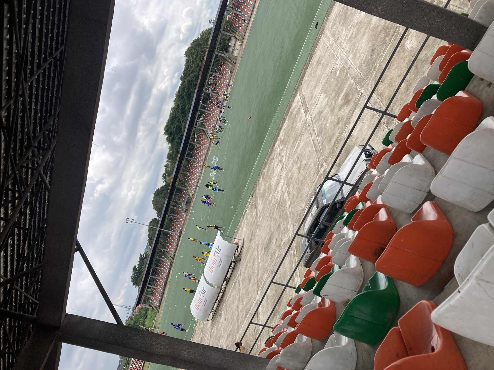 This is a medium shot taken from the stands of the Nest of Champions stadium in Uyo, Nigeria. The stadium features a green football field with several players in athletic attire, engaged in what appears to be a training session. Two white, tunnel-like structures with "Ibom Air" branding are positioned on the edge of the field. The stands themselves are partially visible, with rows of orange, green, and white plastic seats. A dark-colored sedan is parked on the concrete area beside the stands. The sky above is overcast with scattered clouds. The overall scene depicts a sports venue during a practice or training session.