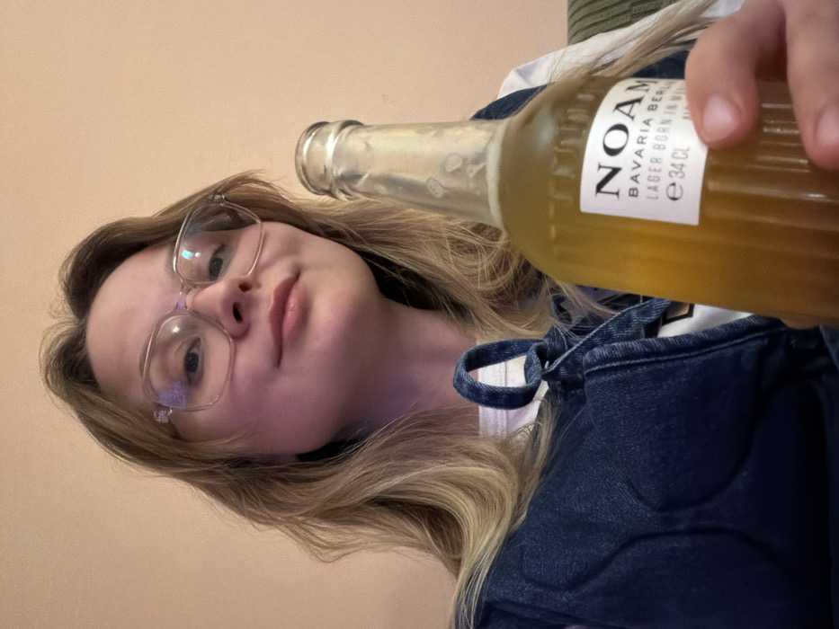 A female subject, appearing to be a young adult, is depicted from a low-angle, close-up perspective in an indoor setting. She has long, light-colored hair and wears clear-framed eyeglasses. Her lips are light-colored, and she looks directly at the camera with a subtle smirk. She is dressed in a dark blue denim overall or jumper layered over a white top. Her right hand holds a clear glass bottle, partially obscuring its lower white label. The bottle contains a light amber liquid, consistent with beer, and is approximately three-quarters full. The visible label text reads "NOAM", "BAVARIA BERLIN", "LAGER BORN IN BERLIN", and "e 34 CL". The background consists of a plain, light peach or orange-toned wall. A small portion of a textured, possibly green-striped upholstered object is visible on the far right. The specific location within the city of Tel-Aviv, Israel, cannot be determined from the visual evidence presented in the image.