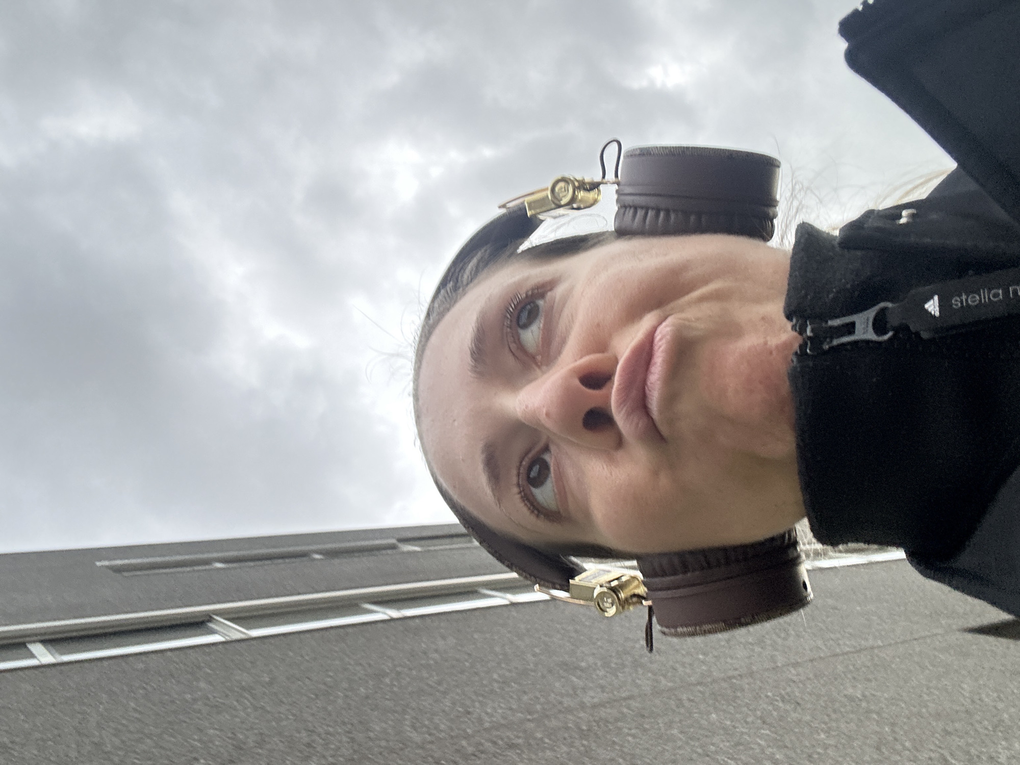 The image is an upward-angled selfie of a woman with light skin and brown headphones. She is wearing a black jacket with a "stella mccartney" label. The sky is cloudy and grey. In the background is a grey building with horizontal white lines, likely windows. The location is possibly Amsterdam, Netherlands. No specific actions or interactions are discernible.
