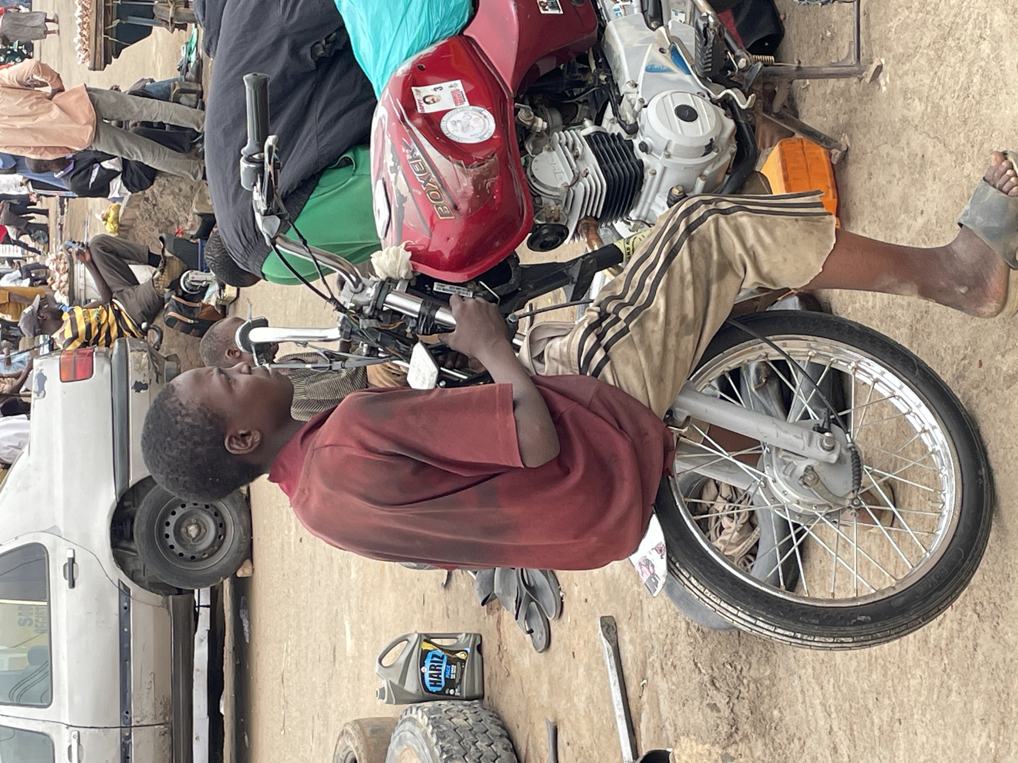 The image captures an outdoor scene, likely a bustling street or market in Jos, Nigeria, during bright daylight hours. The central focus is a young Black male, appearing to be a boy or young man, wearing a red short-sleeved shirt and light-colored pants with dark stripes. He is seated on a red motorcycle, with his hands positioned near the handlebars and engine, suggesting he might be working on it or resting.
The motorcycle is a Bajaj Boxer, with "BOXER" prominently displayed on its red fuel tank, along with a "BAJAJ" logo partially visible and several stickers, including a small portrait photo. Its engine components are visible, showcasing silver and black parts. The front wheel, with its spokes, is also clearly seen.
Around the main subject, the ground is dusty and unpaved, a light brown color. Various items are scattered on the ground, including a black plastic container of engine oil with the brand "HARIZ" visible, an old tire, and several tools, such as a metal lever. A pair of black flip-flops lies near the oil container.
In the background, several other people are present, some standing and others sitting. One person wears a distinctive yellow and black striped shirt. To the left, part of a white car, possibly a van, is visible, with its rear wheel elevated. The overall environment suggests a vibrant, functional area, possibly an informal repair shop or a transit point within a market setting. The atmosphere appears busy and typical of daily life in the region.