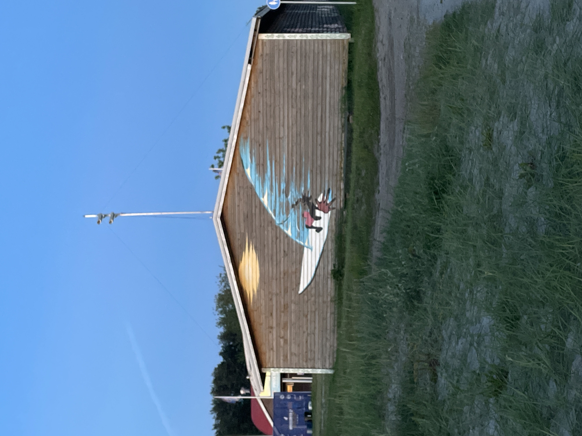 The image shows a mural of a windsurfer on the side of a wooden building, possibly a beach house or a watersports facility. The mural depicts a person on a surfboard with a sail, catching the wind, which is represented by blue streaks trailing behind the sail. The sun is also depicted in the mural as a yellow oval. The building has horizontal wooden planks and a white trim along the roof. To the left of the building, there is a red-roofed structure and some trees. To the right of the mural, a blue and white signpost with a circular blue symbol is visible. The foreground consists of sand and green grass. The sky is clear and blue, suggesting it is daytime. The scene is located in Tallinn, Estonia.