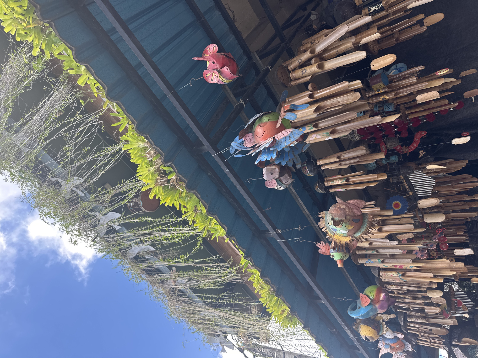The image shows an outdoor stall in Ubud, Indonesia, decorated with various colorful wind chimes and hanging ornaments. The stall has a blue corrugated metal roof supported by a black metal frame. Lush green plants hang from the upper edge of the roof, cascading downwards. The wind chimes are made of bamboo tubes and some have decorative elements like painted flowers or wooden tags. Among the chimes, several whimsical figures are suspended, including a red elephant, a colorful bird with blue wings, a fish, and other animal-shaped ornaments. The scene is bright and airy, with a clear blue sky and white clouds visible in the background. The overall impression is one of a vibrant and artistic marketplace typical of Ubud.