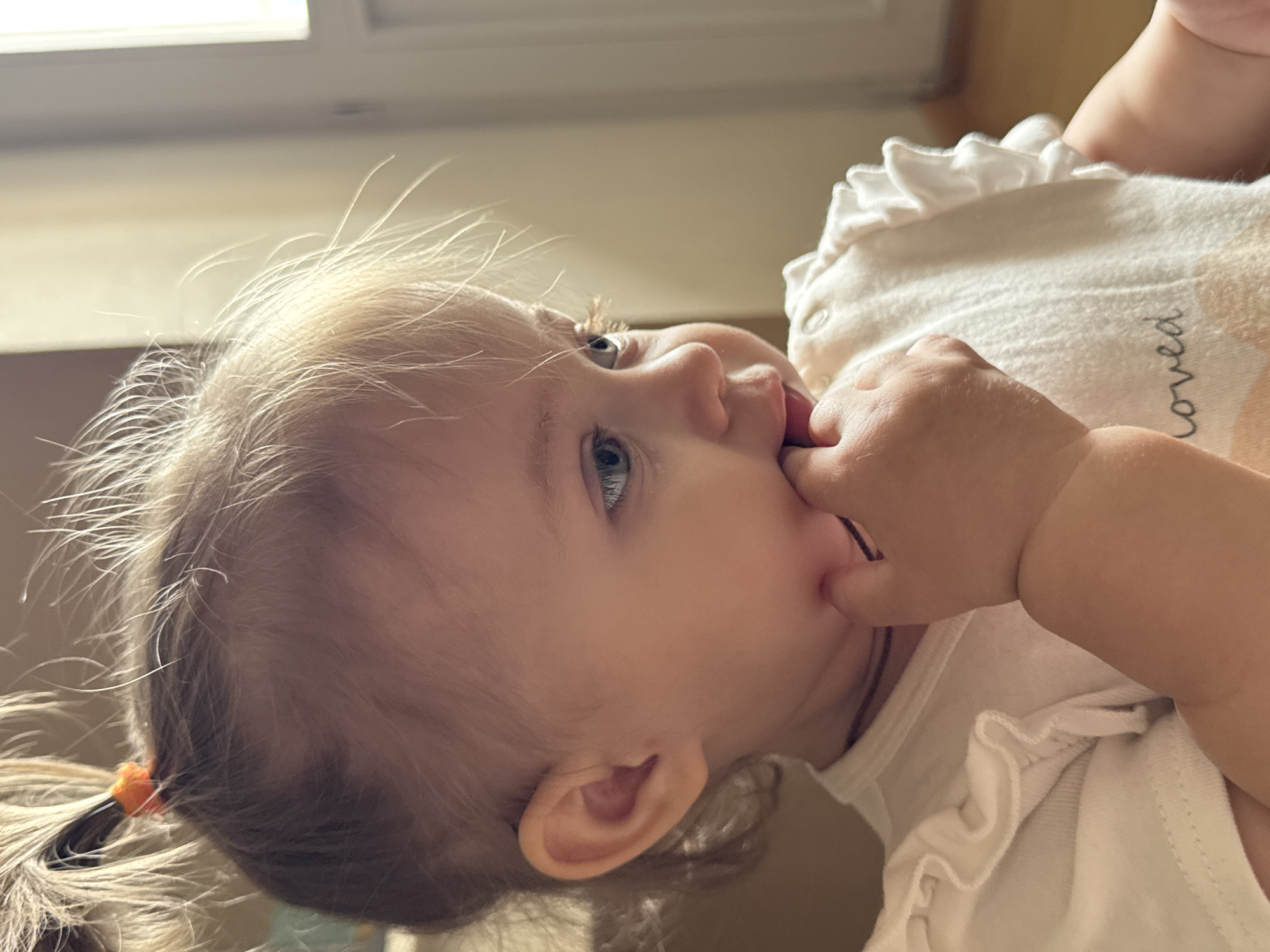 A close-up, vertical, eye-level shot shows the profile of a blonde baby girl. The baby, dressed in a white ruffled shirt with the word "loved" printed on it, is facing right and has her right hand in her mouth. Her blonde hair is pulled into two pigtails with orange elastics. The background is blurry and appears to be indoors, with a door or window visible on the right. The scene is located in Chaikovsky, Russia.