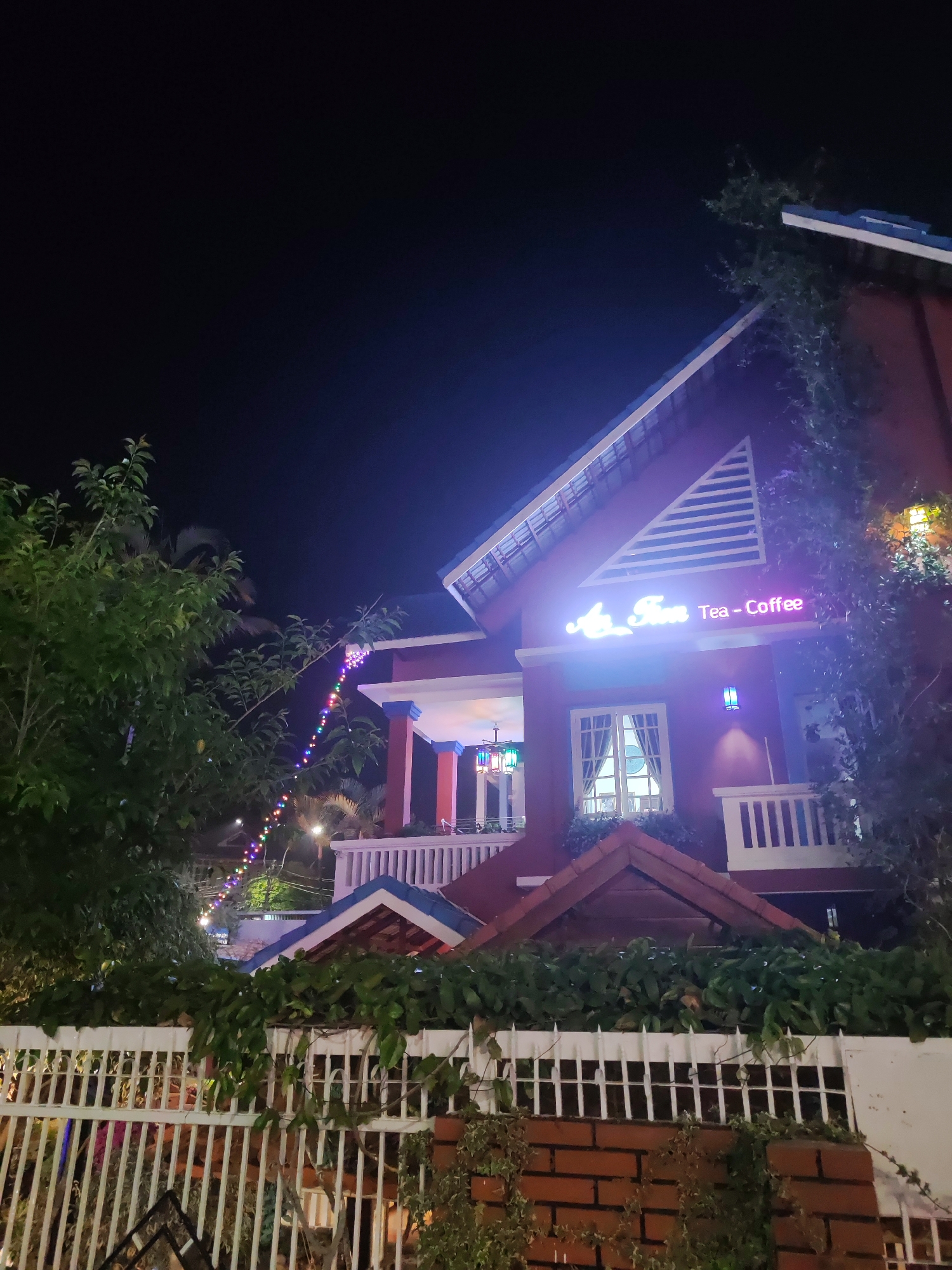 The image shows a two-story building at night, identified by a neon sign as "Ai Tiem Tea - Coffee." The building is painted in shades of red and is trimmed with blue. There's a white picket fence in the foreground, partially obscured by vegetation. String lights are visible in the background, extending over the fence. The image was likely taken in Da Lat, Vietnam.