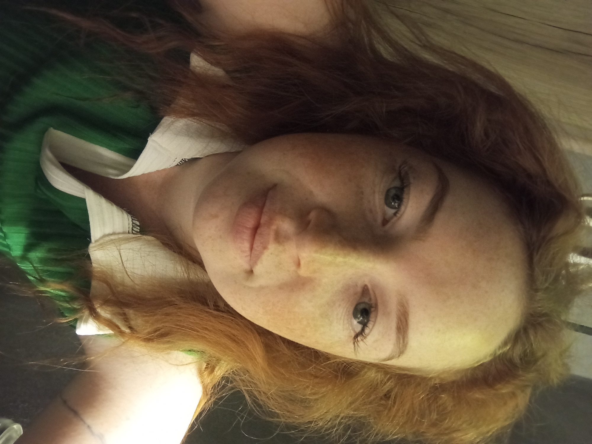 A selfie shows a fair-skinned woman with red hair and many freckles on her face. She has light-colored eyes and a neutral expression. She is wearing a green shirt with a white collar. Part of her left arm is visible, with a faint tattoo on the inner forearm. The background appears to be a dark, possibly wooden, surface. There is no information available in the image regarding the location as Yurgamysh, Russia.