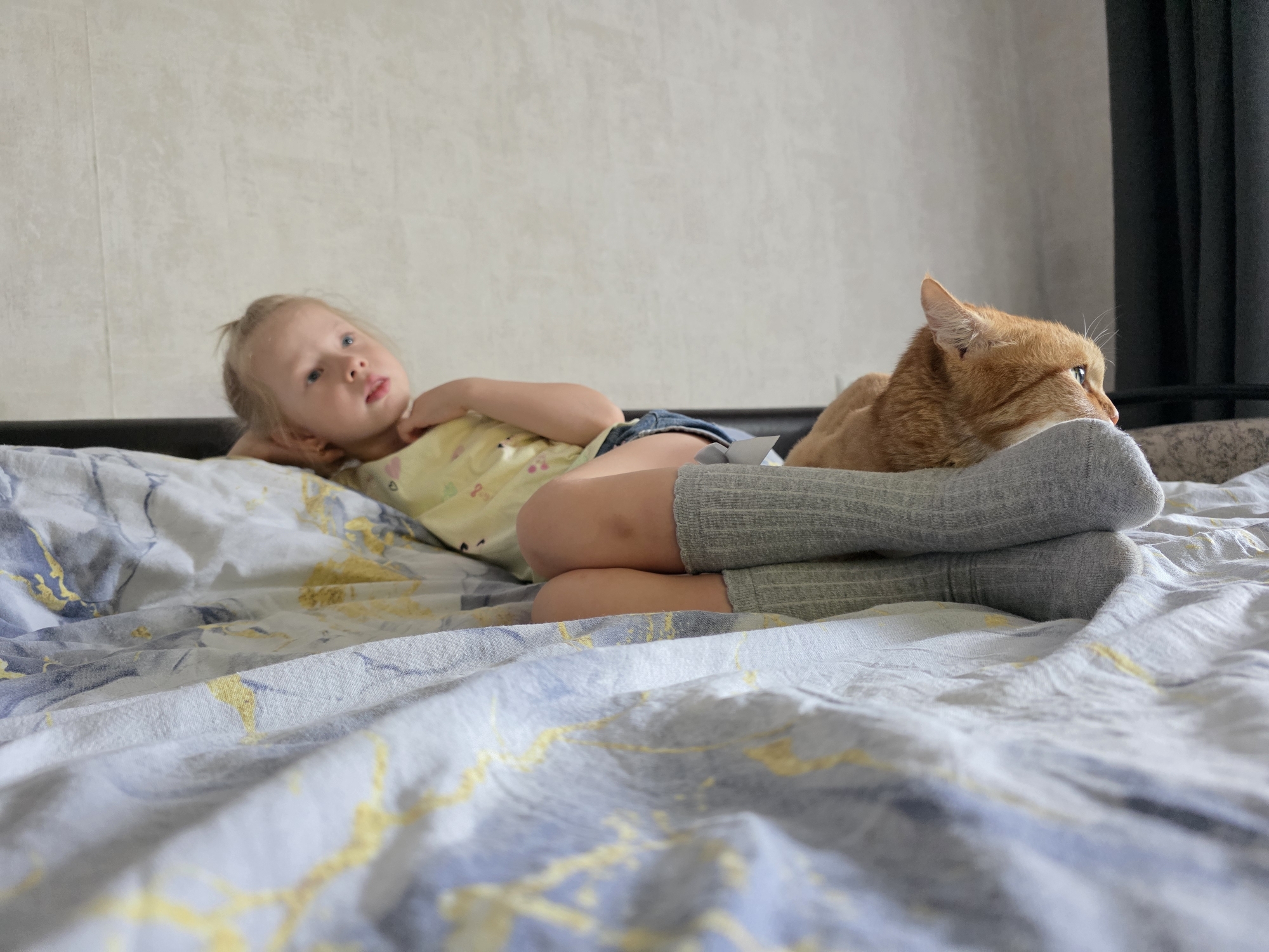 A young girl with blonde hair and blue eyes is lying on a bed with a cat. The girl is wearing a yellow shirt with a floral print and denim shorts, and grey knee-high socks. She is propped up on her left arm, looking away from the camera. Her legs are bent, with the right leg resting on top of the left. A ginger cat is lying next to her, with its head resting on her leg. The cat is looking to the right, with its eyes partially closed. The bed is covered with a light-colored duvet with a blue and gold marbled pattern. In the background, a plain grey wall and a dark curtain are visible. The lighting suggests it is daytime. The scene appears to be indoors, likely in a bedroom. There is no visible text in the image.