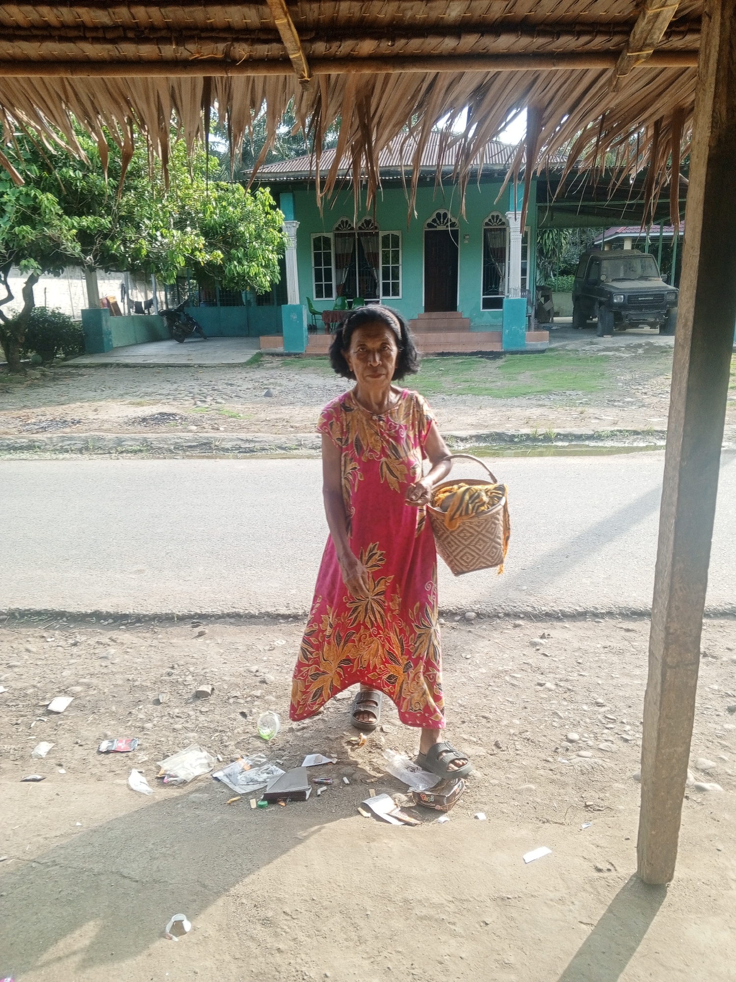 A woman stands on a dirt patch outside a building in Tanah Gara Hulu, Indonesia. She is wearing a pink dress with a yellow floral pattern and carrying a woven basket in her right hand. The building behind her is light blue with white trim and arched windows. A black SUV is parked to the right of the building. The ground is covered in dirt, gravel, and scattered litter, including plastic bottles, wrappers, and paper. A thatched roof overhang is visible in the foreground, casting a shadow on the ground.