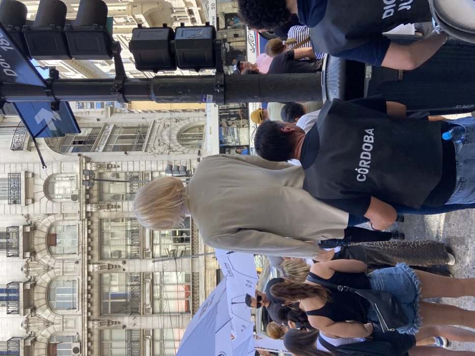 This image depicts an outdoor urban scene occurring in the Microcentro district of Buenos Aires, Argentina. The central focus is the back of a remarkably tall figure with short blonde hair, wearing a light beige blazer over blue jeans. This figure's unusual height suggests it is either a puppet, a person on stilts, or a similar elevated construct, towering over the surrounding individuals.
Immediately in the foreground, to the right, a man is seen from behind, wearing a black t-shirt and a black vest bearing the white capital letters "CÓRDOBA" on its back. A blue plastic water bottle is visible in his back pocket. He is positioned closely behind the tall blonde figure, appearing to be an attendant or operator.
A diverse crowd of people occupies the middle ground and left side of the frame. To the left, several individuals are gathered under white umbrellas, some displaying red logos. One man under an umbrella wears sunglasses. A woman with dark hair in a ponytail, wearing a black tank top and denim shorts with a black fanny pack, is prominent on the left.
The background is dominated by a grand, multi-story building featuring ornate, light-colored European-style architecture with multiple arched windows, balconies, and decorative stonework. On the right, a black utility pole supports a vertical traffic light assembly (lights appear amber/off) and a blue street sign with a white left-pointing arrow and the numbers "1800". A dark waste receptacle or utility box is attached to the base of the pole. A white and blue public bus, marked "Central" and "50," is visible in the middle ground behind the crowd, indicating a busy urban thoroughfare.