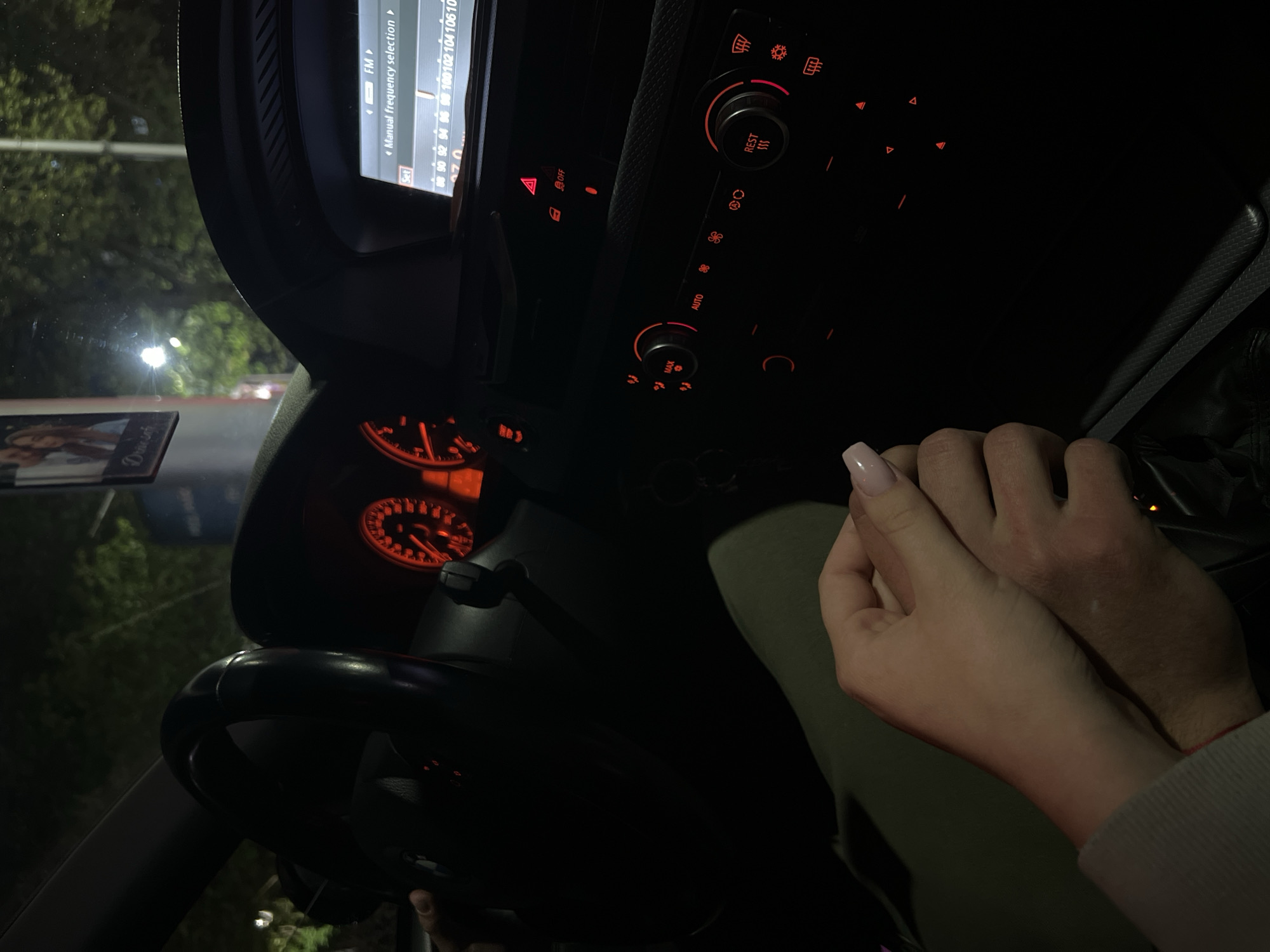 The image depicts the interior of a car at night, viewed from an upward angle and rotated counter-clockwise. In the foreground on the right, two hands are clasped together, with a lighter-skinned hand (featuring light pink manicured nails) holding a darker-skinned hand with visible arm hair, suggesting intimacy or connection.
The car's dashboard is prominent, illuminated by glowing red and orange lights. A central infotainment screen displays radio settings, including "FM" and "Manual frequency selection," along with a frequency band showing "90 92 94 96 98 100 102 104 106 10". Below this, the climate control panel features various buttons such as "OFF," "MAX," "AUTO," and "REST," along with hazard lights and fan controls, all lit in red. To the left, a portion of the instrument cluster, likely a speedometer or tachometer, also glows with a reddish-orange light. Part of the steering wheel is visible in the lower left.
Through the windshield, the dark forms of trees and distant artificial lights are visible against the night sky, indicating it is late evening or night. A small card or photo on the dashboard, partially obscured, shows two people and the word "Drive." The scene captures a quiet, personal moment within the vehicle, likely in Plovdiv, Bulgaria, during an evening journey or stop.