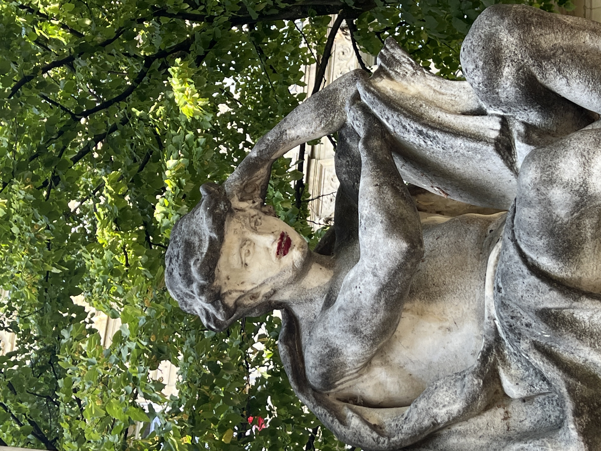 The image is a close-up shot of a weathered, grey stone sculpture of a male figure. The sculpture is in a seated or reclining pose, with one arm resting on its head and the other bent at the elbow. The figure appears to be draped in fabric, and there is a distinct application of bright red lipstick on its lips. The sculpture is set against a backdrop of lush green foliage from a tree, with hints of a building visible through the leaves. The lighting suggests it is daytime, with sunlight filtering through the leaves, creating dappled shadows. There are no people or animals present in the immediate vicinity of the sculpture. The overall impression is one of an artistic, albeit somewhat unusual, display in an outdoor urban setting. The sculpture is located in Paris, France, specifically in the Jardin des Tuileries, a public garden situated between the Louvre Museum and the Place de la Concorde.